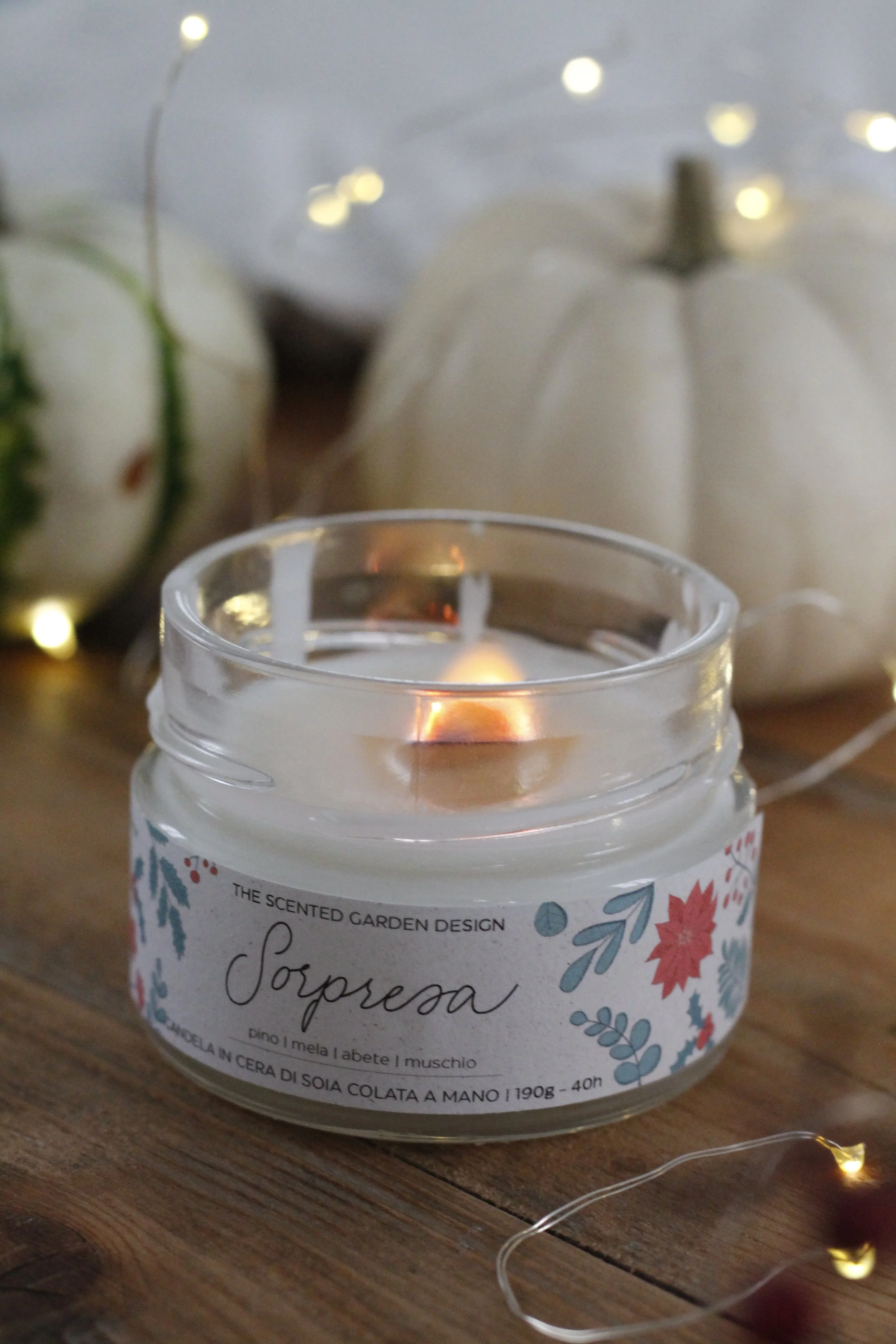 Candea profumata in un barattolo di vetro con etichetta decorativa, circondata da zucche bianche e luci decorative su una superficie di legno.