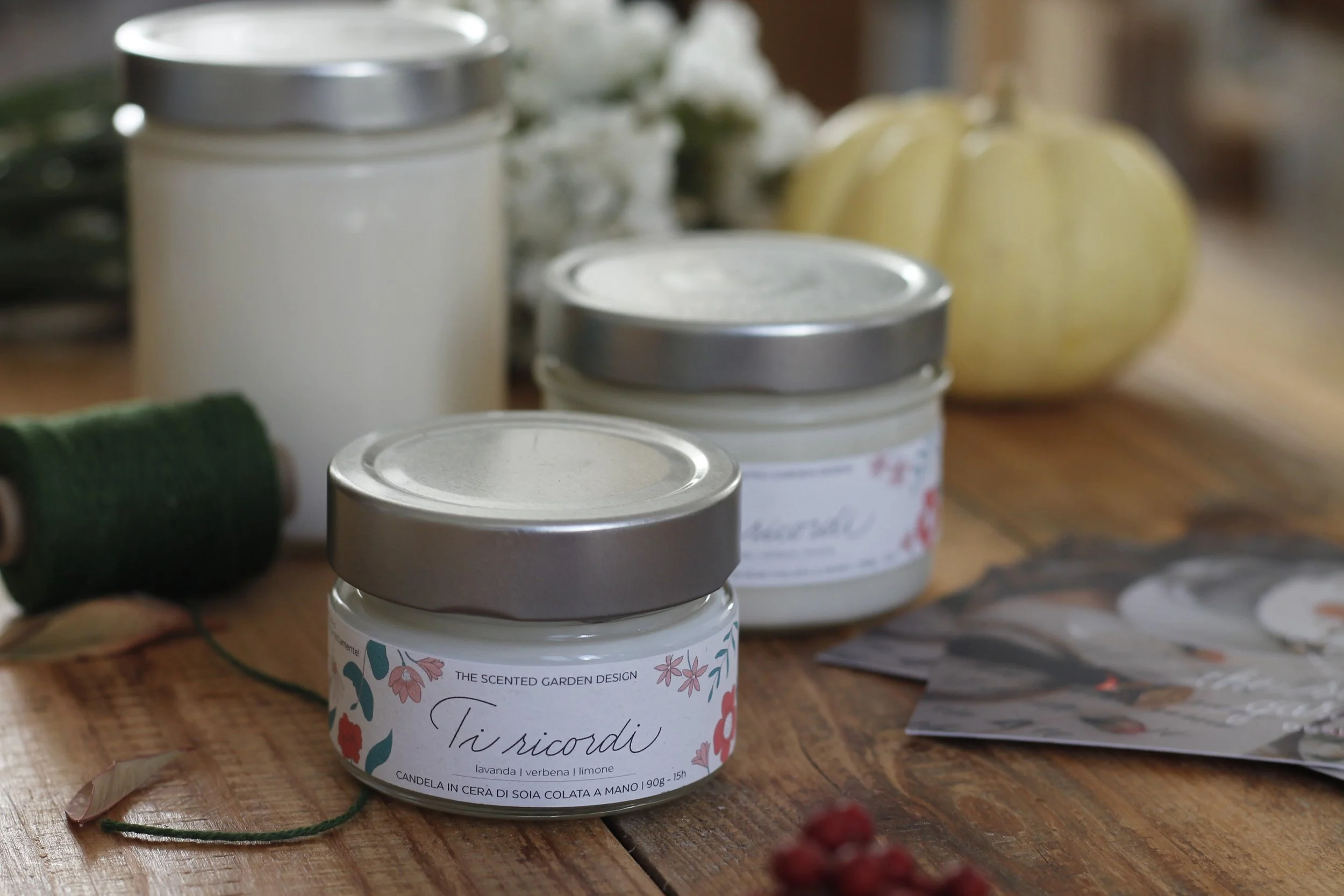 Barattoli di candele artigianali con etichette decorative, presenti su un tavolo di legno, decorato con fiore e bacche.