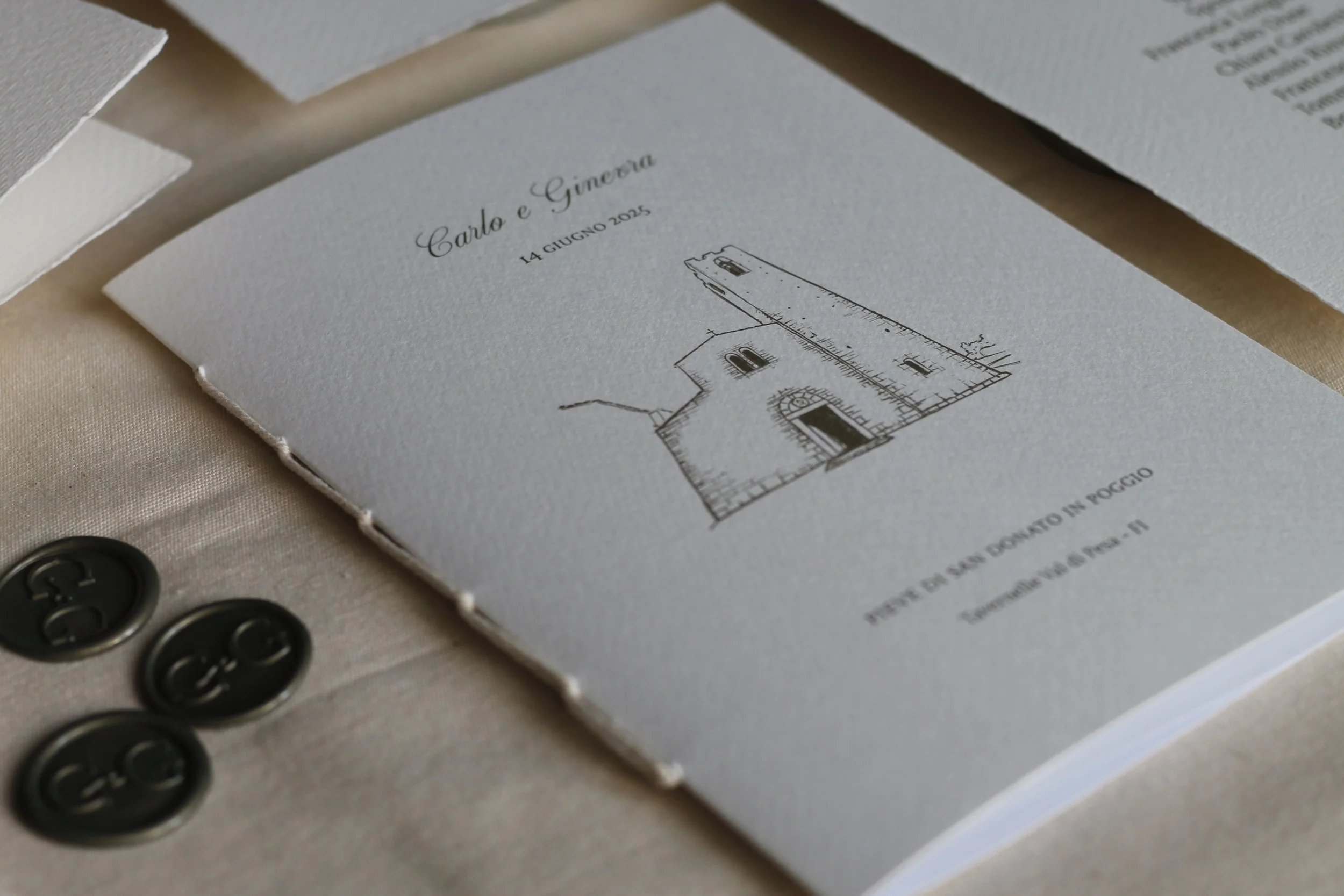 Menu di nozze su carta color avorio con disegno di una torre medievale, con dettagli su data e luogo. Accanto, bottoni neri sulla stoffa coordinata.