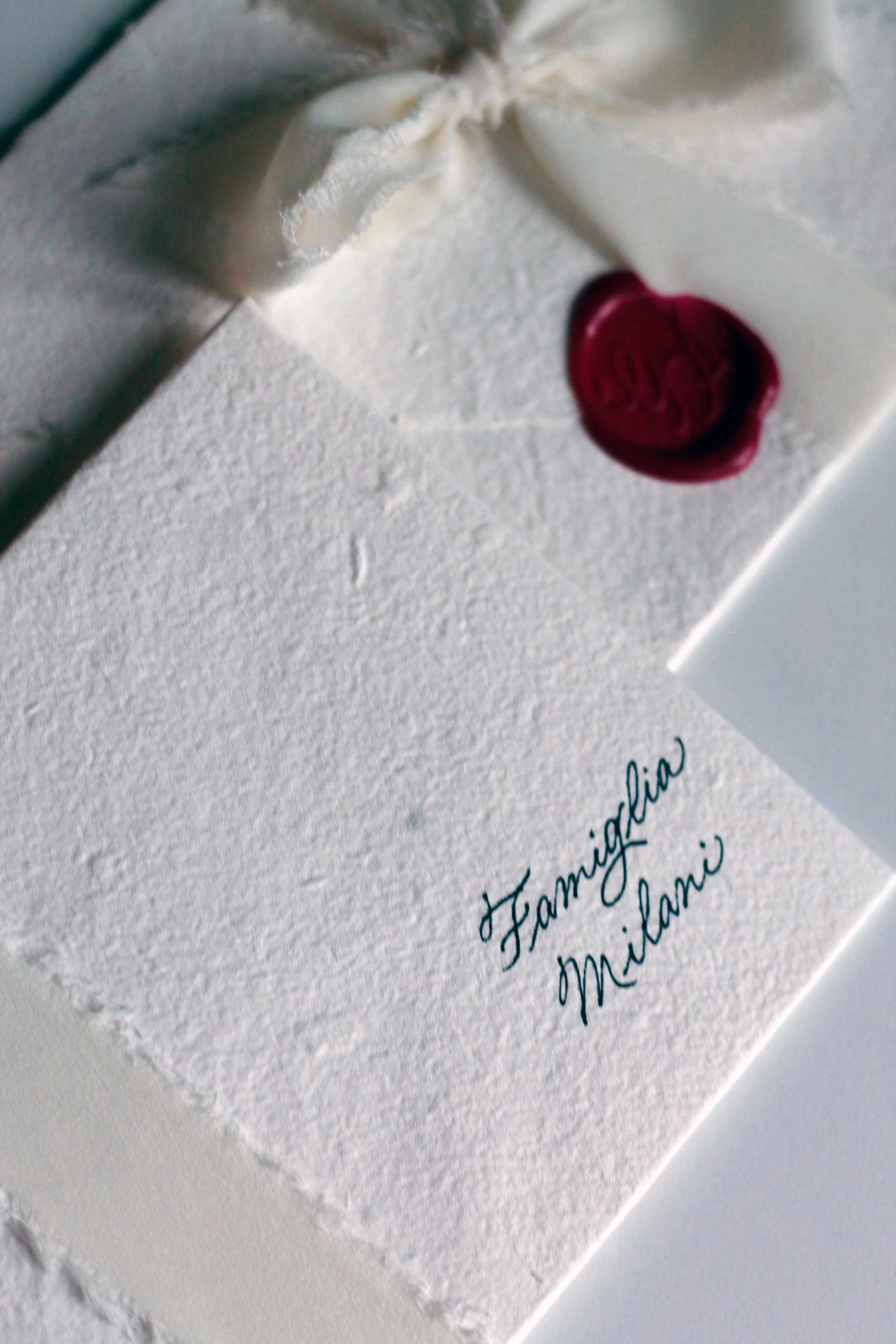 Una busta bianca con un sigillo di cera rossa, una nappina di tessuto bianco e un cartoncino con scritto 'Famiglia Milani' in corsivo.