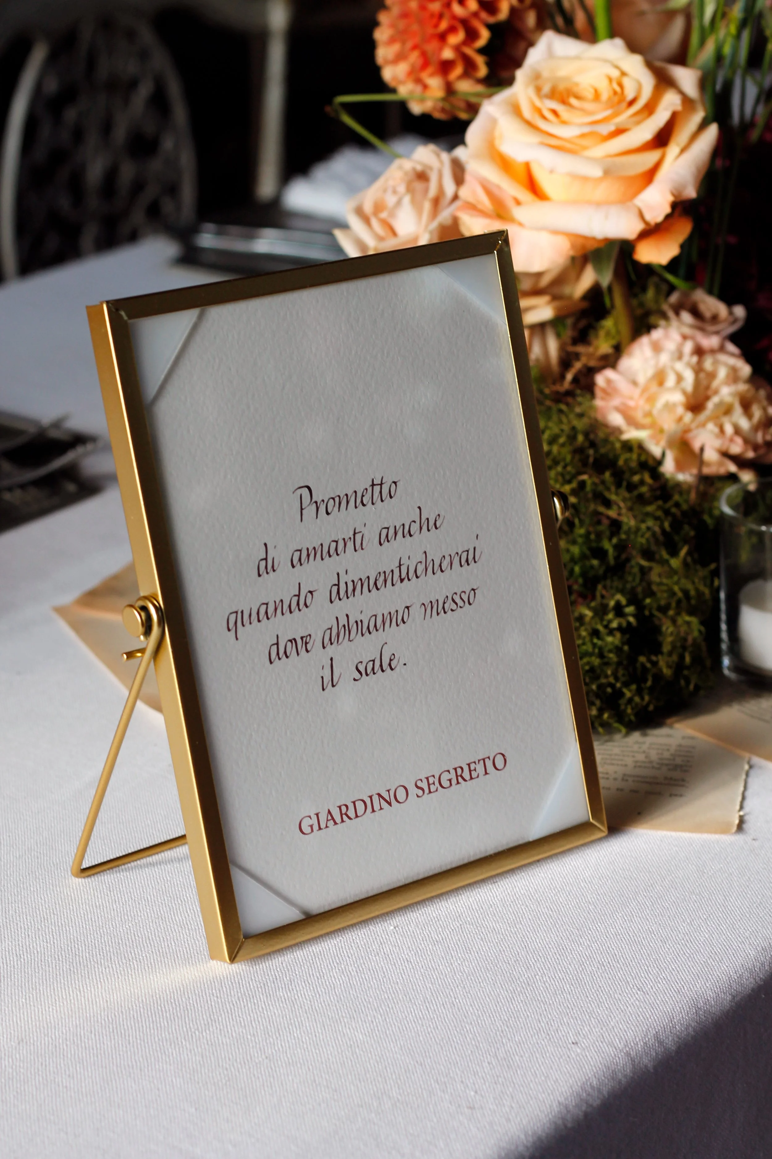 Un tavolo decorato con un quadro con una poesia, fiori rosa e piante verdi sullo sfondo di una tavola apparecchiata per un evento o matrimonio.