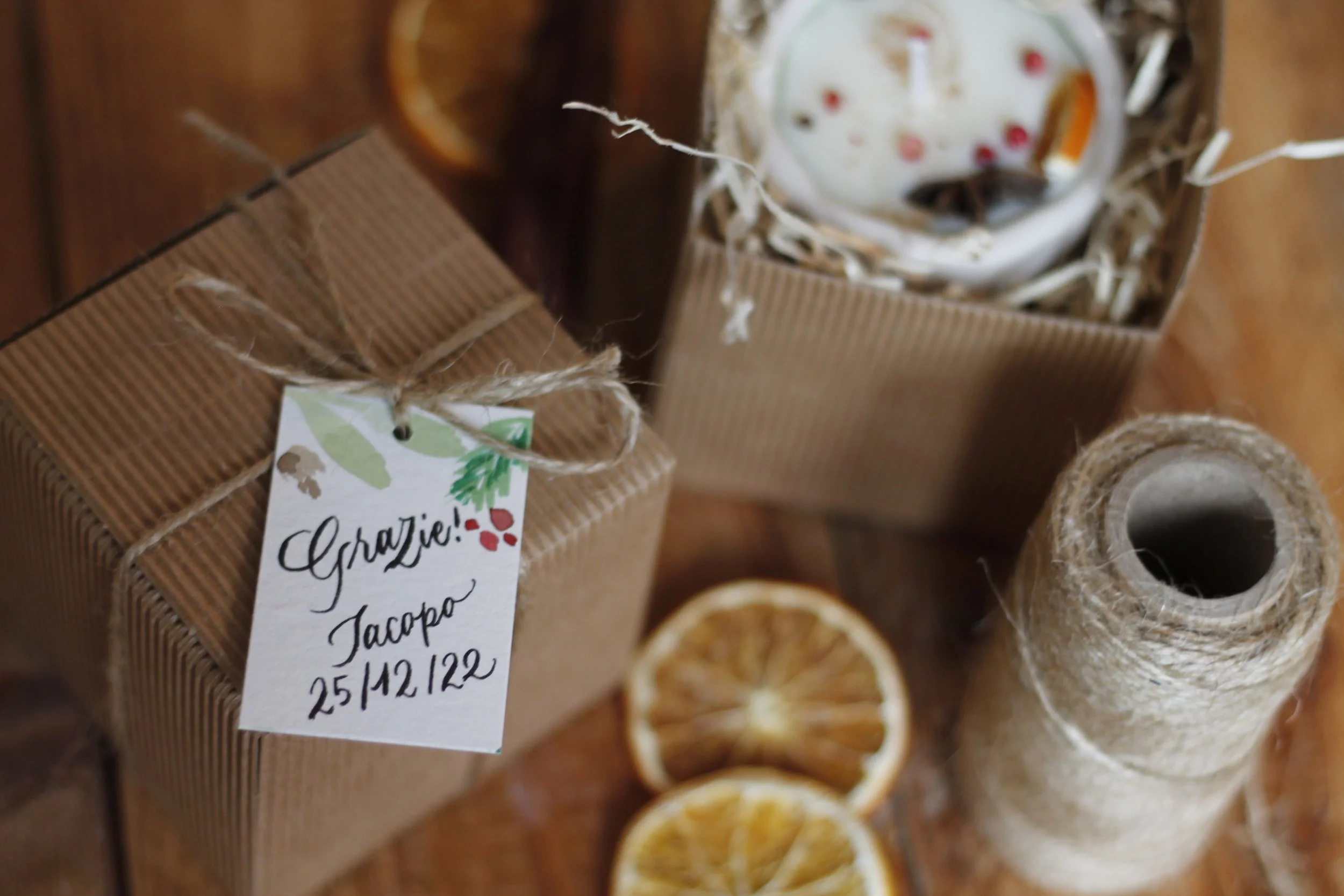 Regalo di Natale incartato con nastro di juta e bigliettino con scritto 'Grazie! Jacopo 25/12/22', palle di Natale, arance secche, fiocco di lana, e una tazza di latte con gocce di ciliegia e spezie.