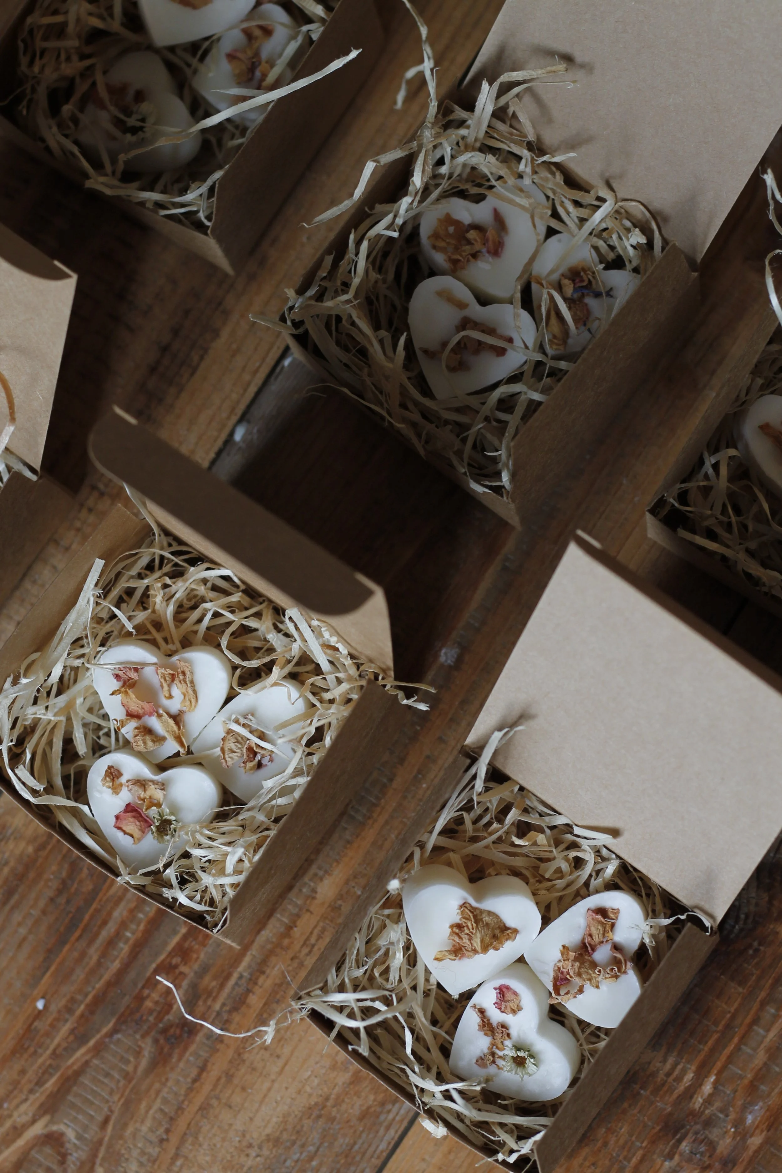 Cuori di cera bianchi decorati con petali di fiore essiccati, sistemati in scatole di cartone con paglia, disposte su un tavolo di legno.