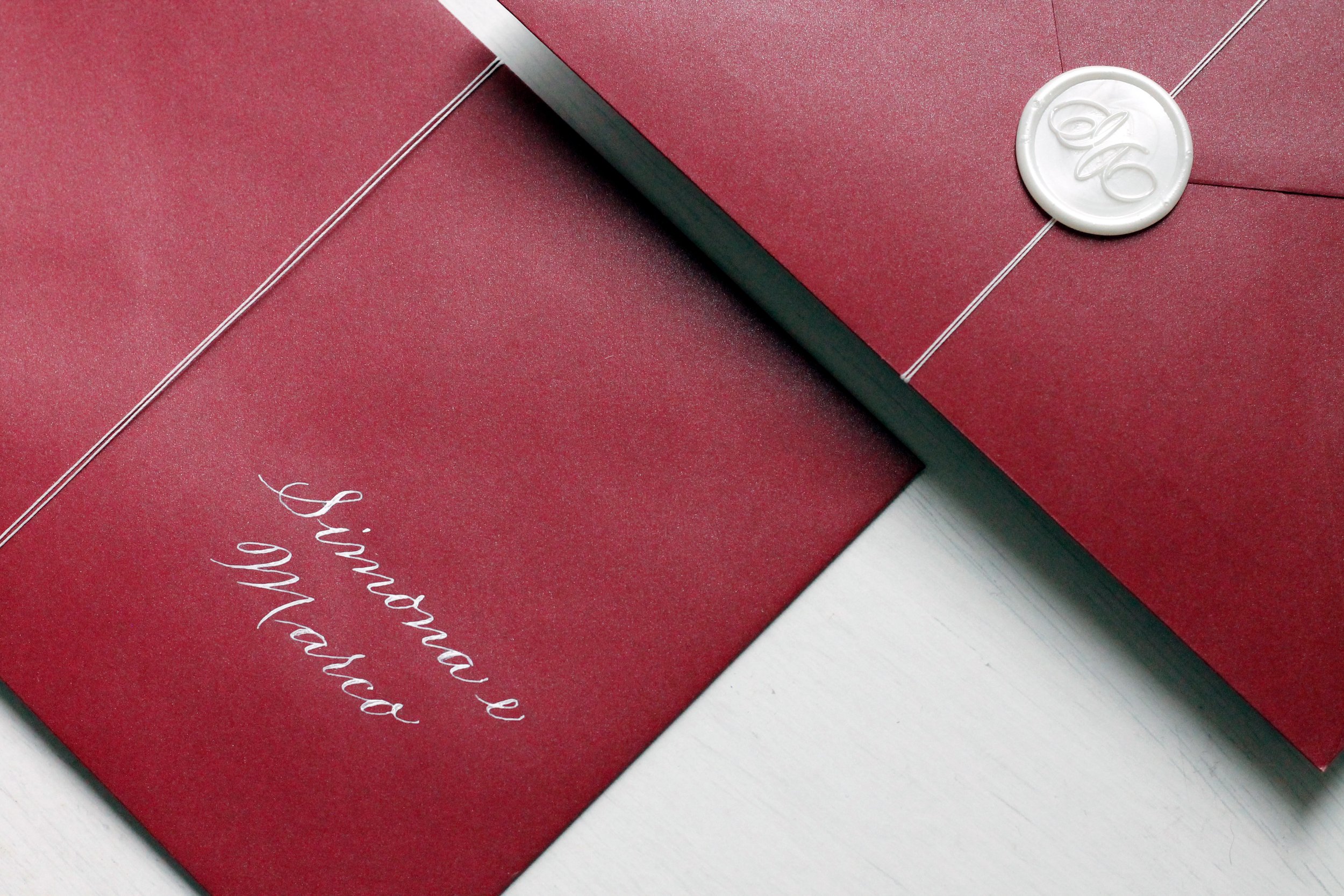 Invito di nozze con testo scritto a mano su sfondo rosso, decorato con ceralacca bianca e filo metallico.