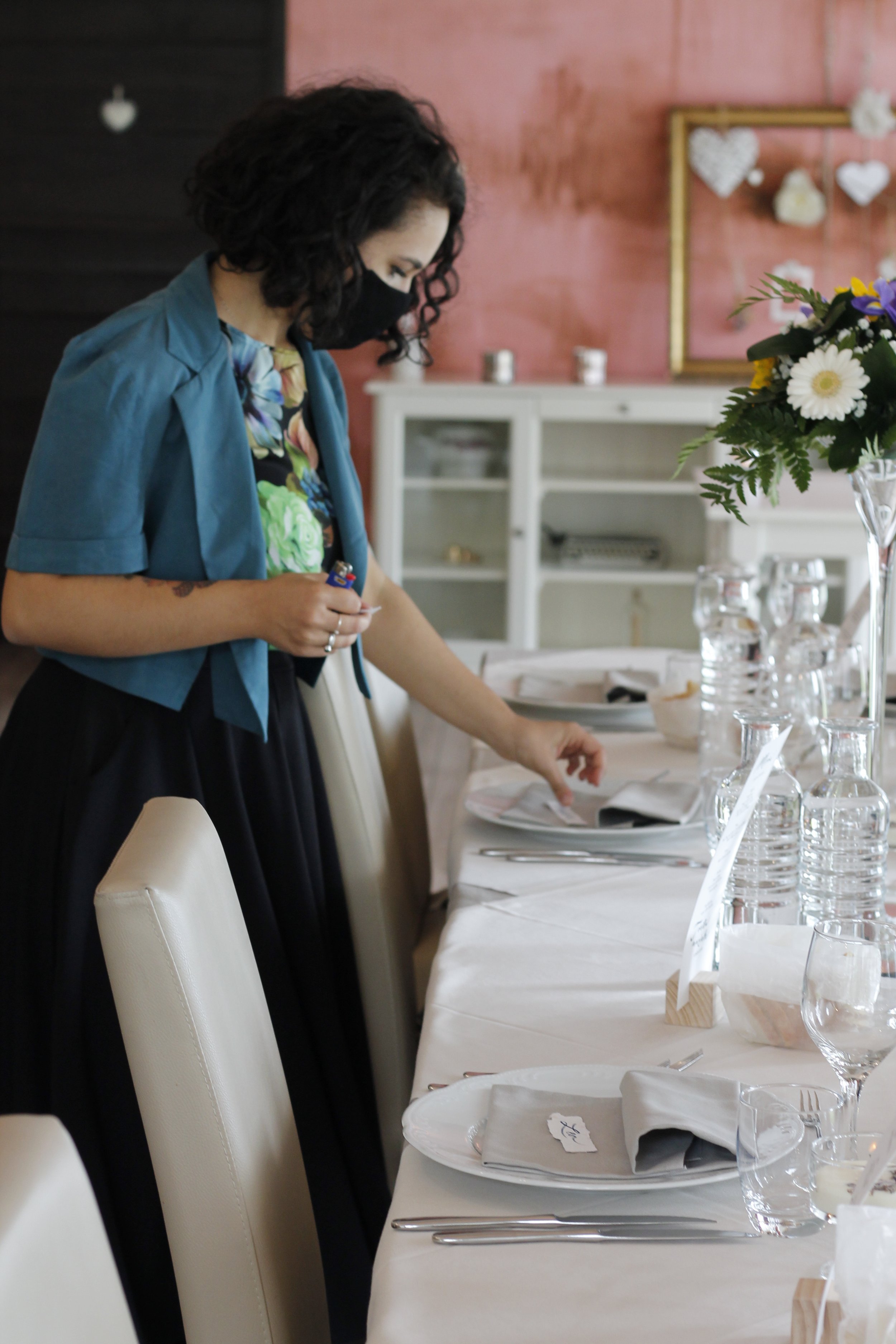 Donna con mascherina che sistema una tavola per un evento o una celebrazione in una stanza decorata con fiori e cuori di carta.