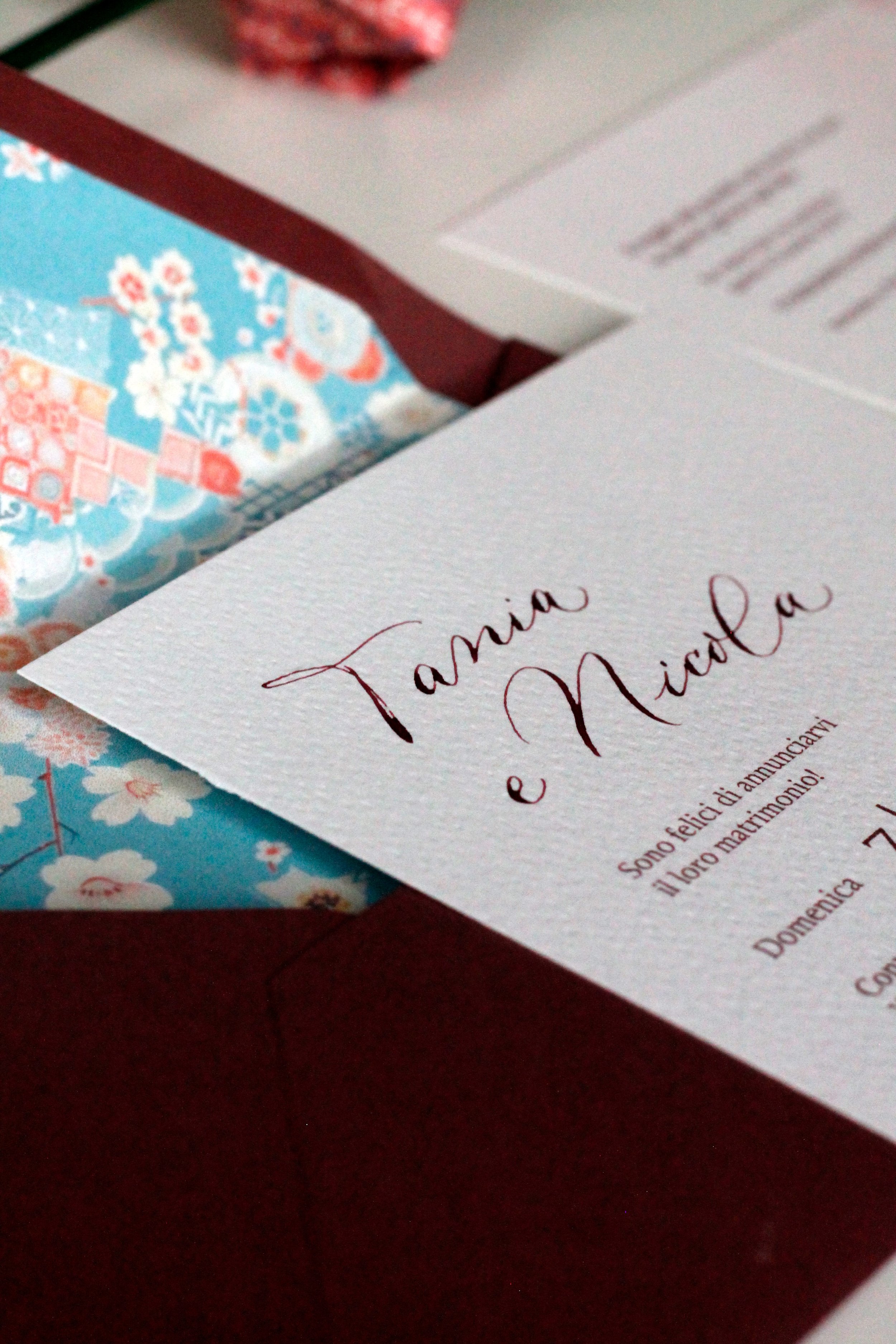 Invito di nozze con scrittura elegante, parzialmente visibile, e tre pezzi di carta colorata con motivi floreali e geometrici, all'interno di una busta rossa.