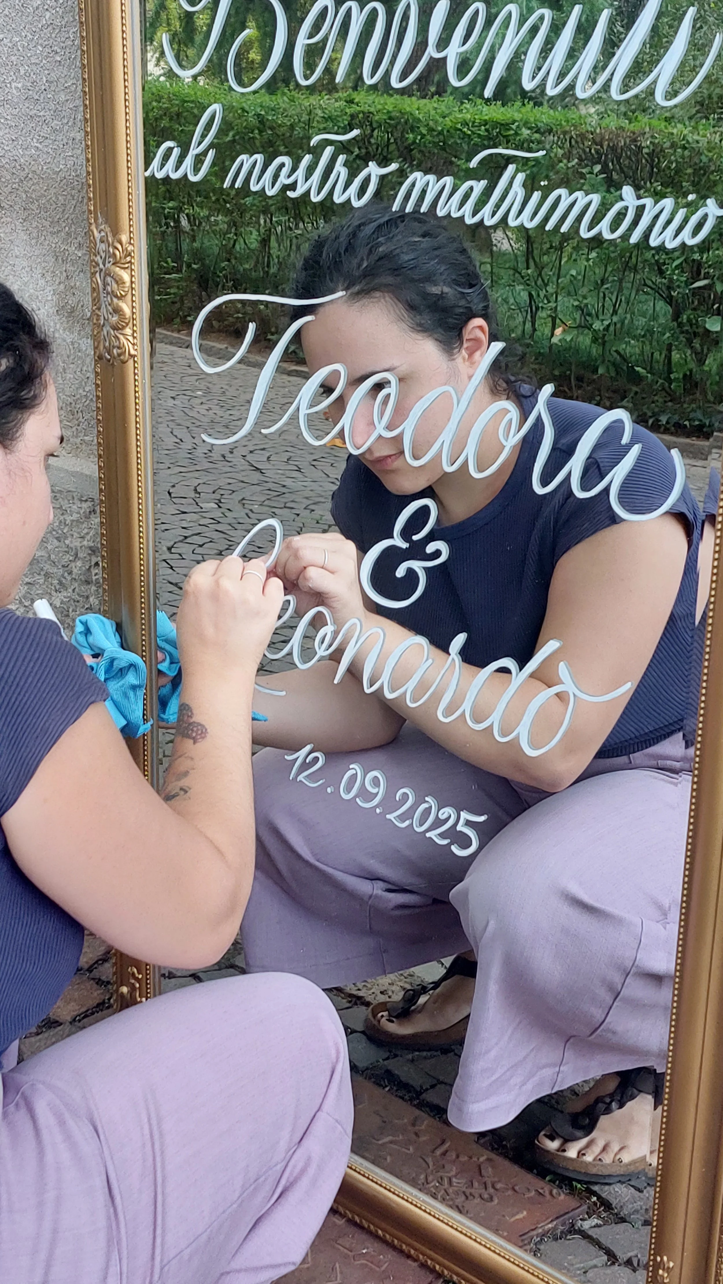 Una donna si guarda allo specchio incorniciato, dove sta scrivendo con un pennarello. Il testo scritto sullo specchio dice: 'Benvenuti al nostro matrimonio Tecido & Eomardo 12.09.2025'. La scena si svolge all'aperto, con un cammino di pietra e del verde sullo sfondo.