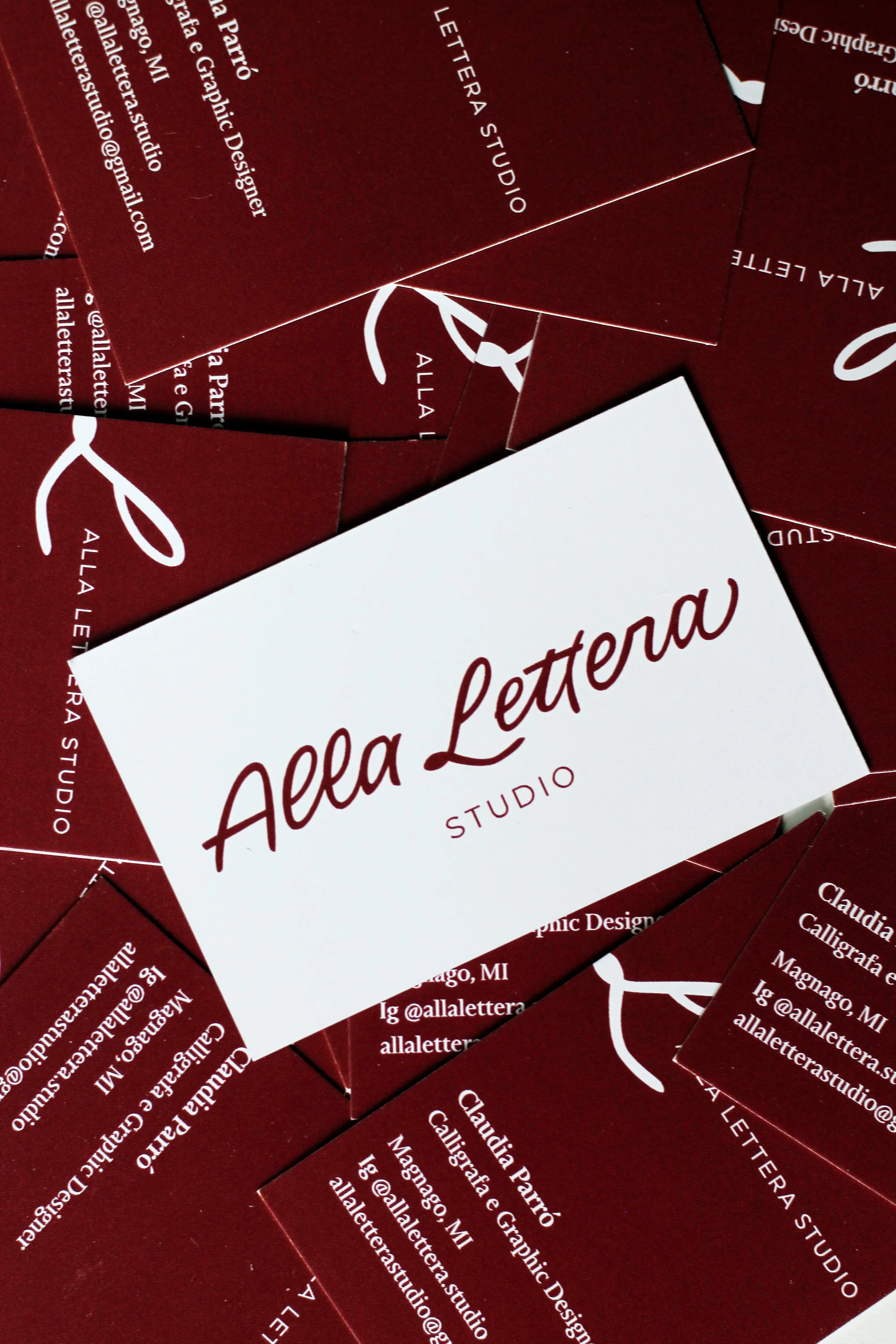 Un mazzo di biglietti da visita con sfondo rosso scuro e testo bianco, mostrando il nome 'Alla Lettera Studio' e informazioni di contatto.