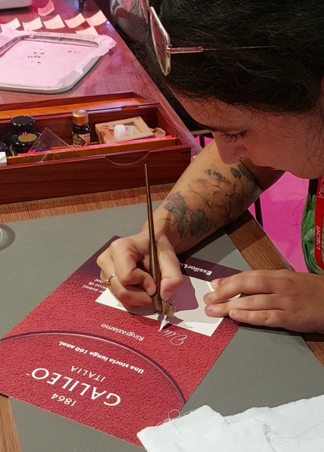 Una persona con tatuaggi sulla braccia sta firmando un certificato di calice su un tavolo di legno con una penna tradizionale.