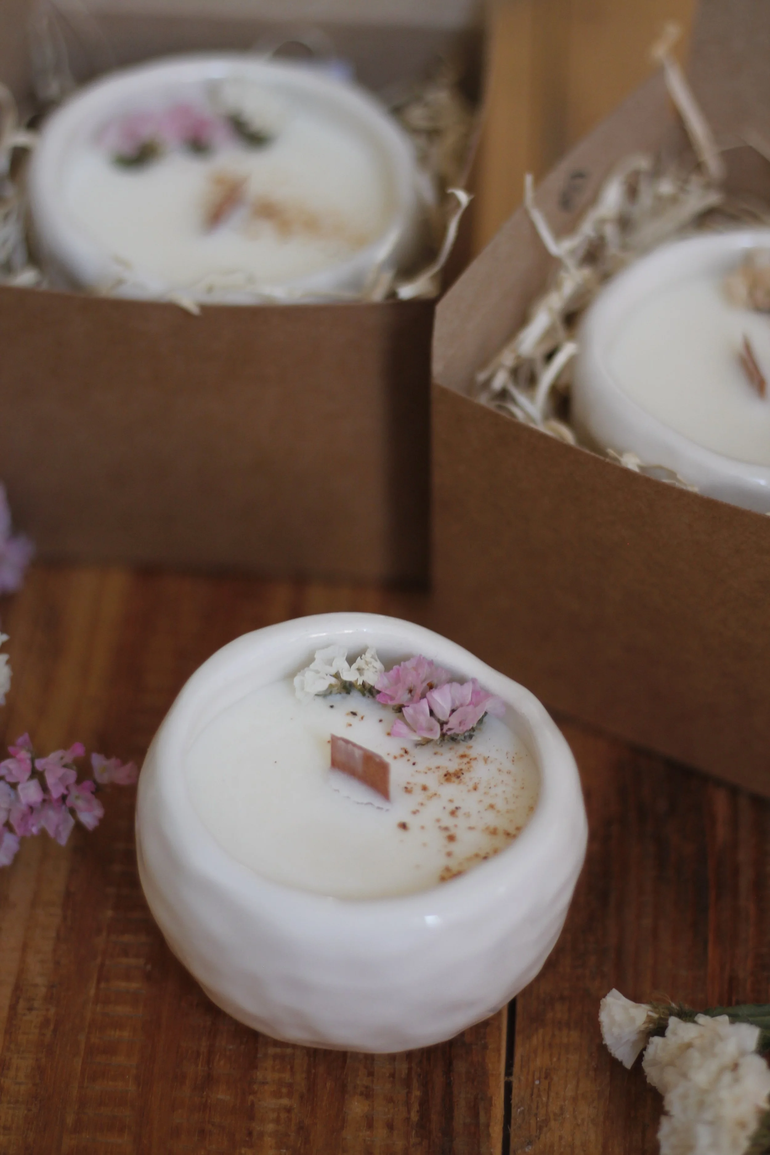 Candele aromatiche bianche decorate con fiori secchi rosa e marroni in ciotole di ceramica, su un tavolo di legno.