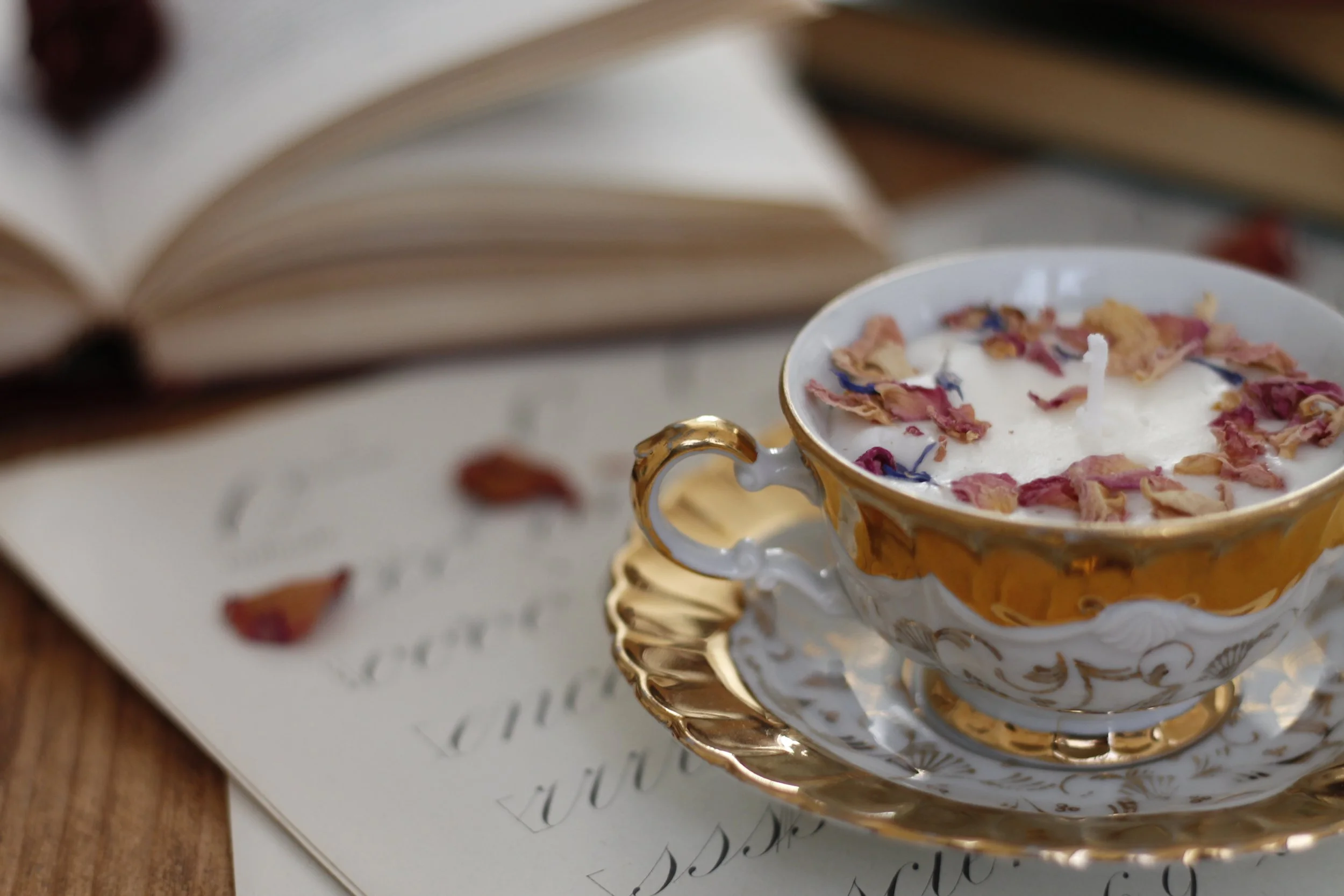 Una tazza di ceramica decorata con motivi dorati, piena di crema o latte con petali di rose sopra, su un piattino dorato. Sullo sfondo un libro aperto e una pagina con scrittura calligrafica.