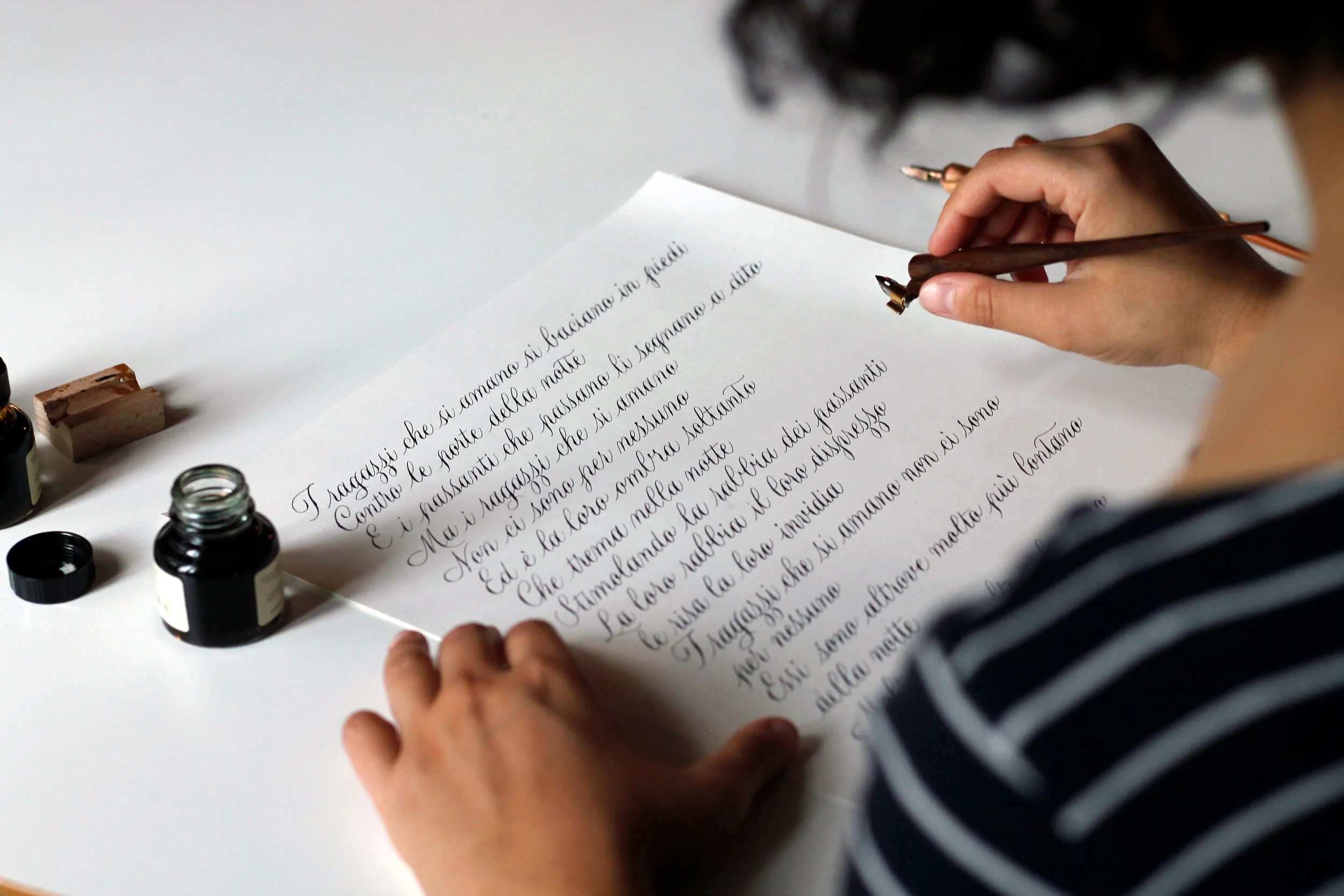 Persona che scrive una poesia con inchiostro nero su un foglio bianco, con bottiglie di inchiostro e pennino sul tavolo.