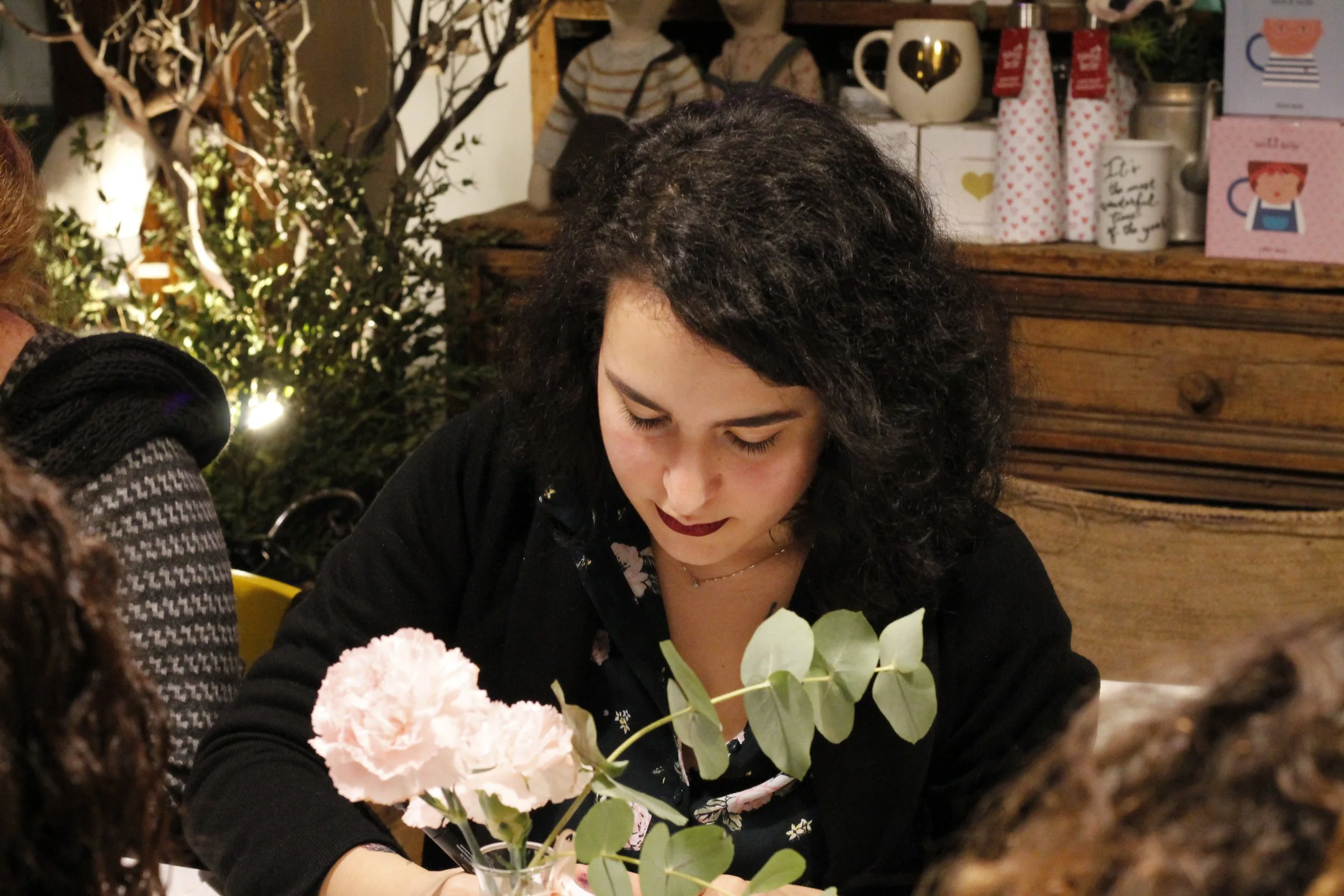 Una donna con i capelli ricci neri, vestita di nero, che guarda con attenzione un bouquet di fiori pastello e foglie verdi su un tavolo in un ambiente decorato con oggetti in cucina, luci calde e persone sullo sfondo.
