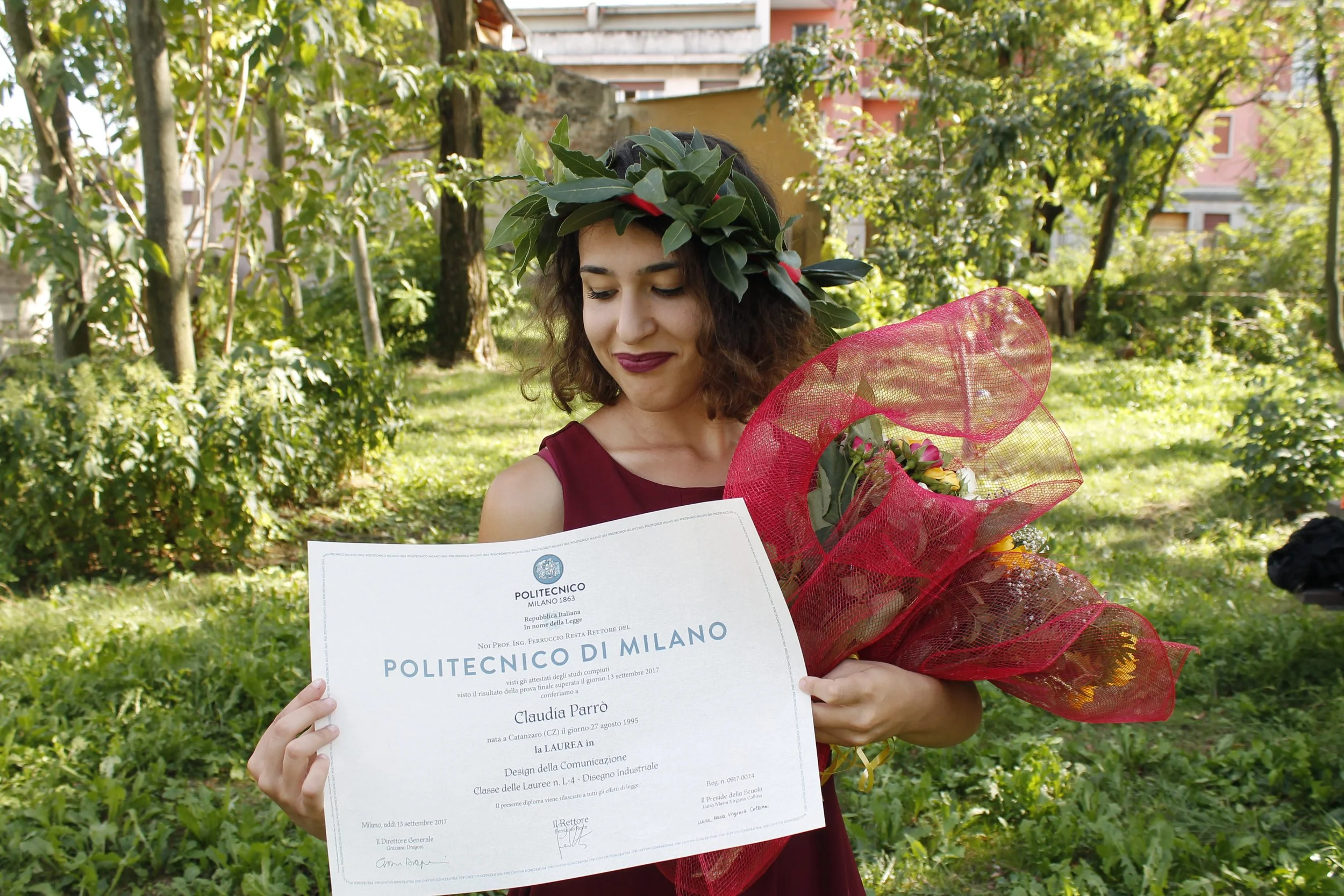Una giovane donna con una corona di foglie e bacche, vestita di rosso, sorride mentre tiene un diploma e un bouquet di fiori in un giardino verde.