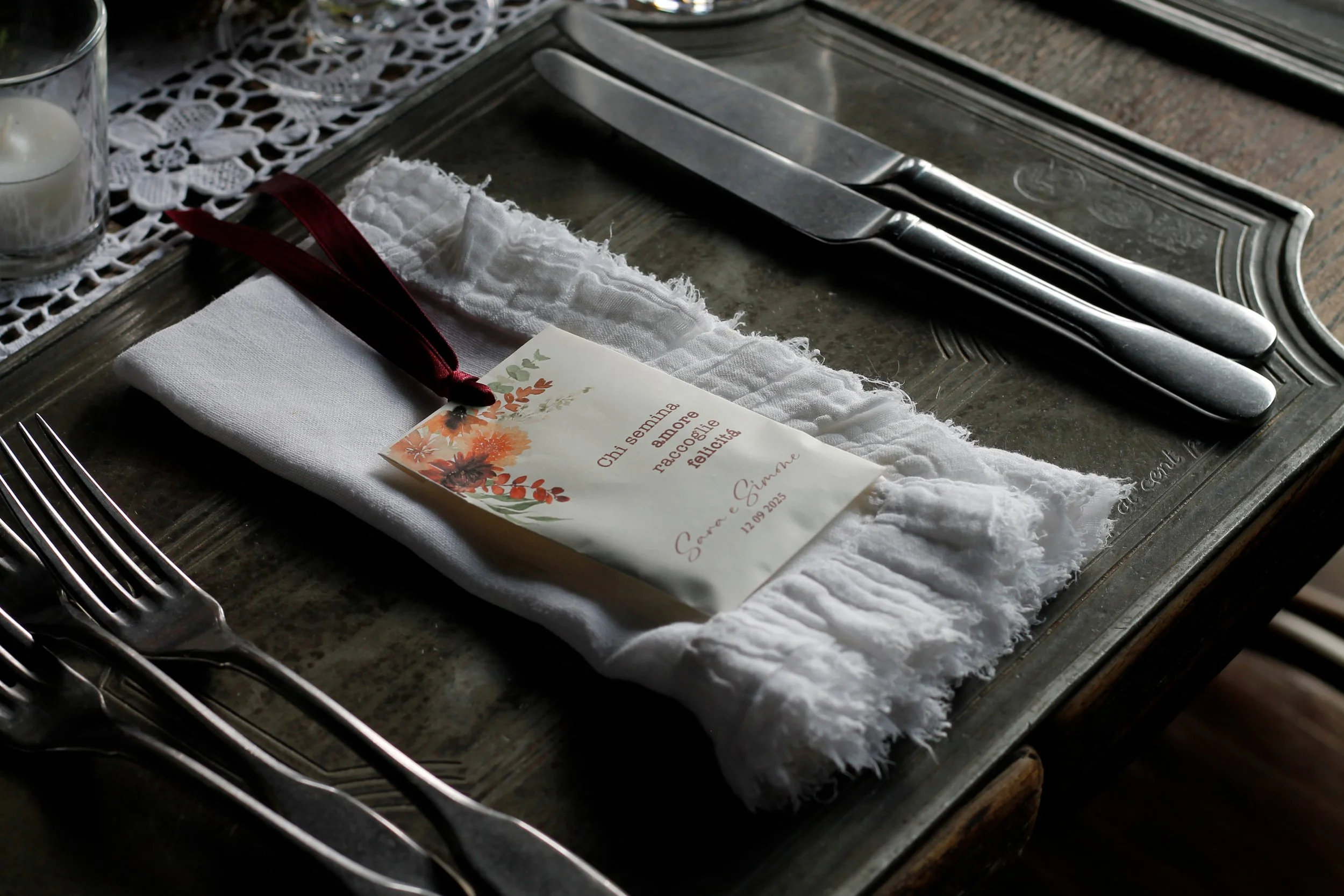 Tavola apparecchiata con coltelli e forchette di metallo, un tovagliolo bianco con bordi frangiati, e uno straccio bianco. Su il tovagliolo c'è un biglietto di ringraziamento con un motivo floreale, indirizzato a persone che hanno condiviso una settimana di amore e felicità, con una firma e la data 12.09.2015.
