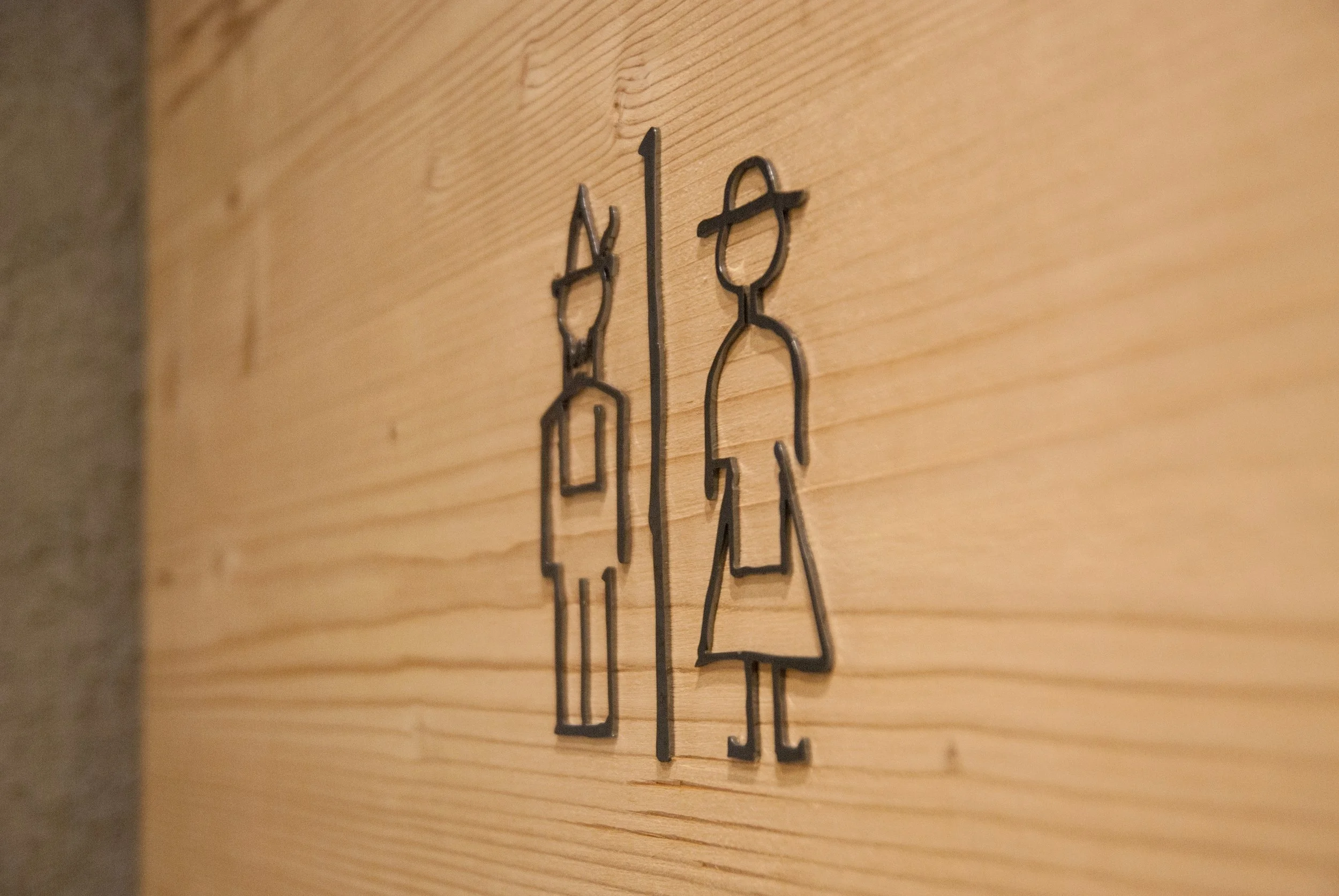Metallene Umrandung eines Damen- und Herrentoilettenzeichens an einer Holzwand.