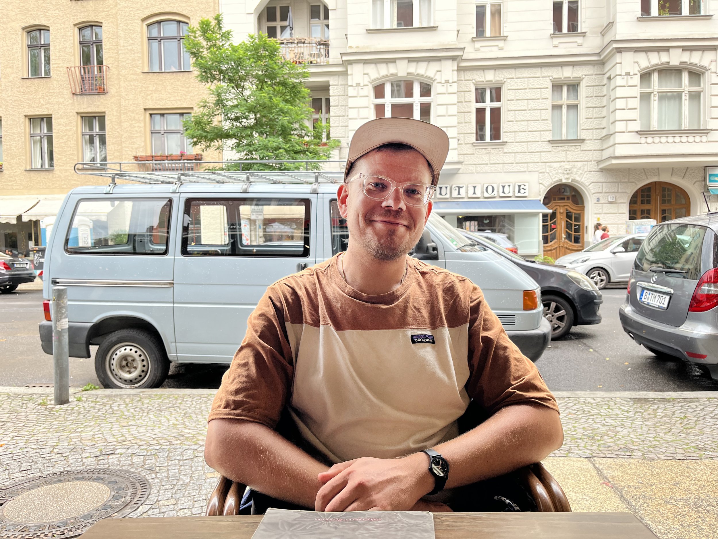 Ein lachender junger Mann, Matthias Krissmayr, sitzt an einem Tisch vor einem Café, trägt eine Basballkappe, Brille, ein beige-braunes T-Shirt und eine Armbanduhr, im Hintergrund sind parkende Autos, ein grauer Van und mehrstöckige Gebäude sichtbar.