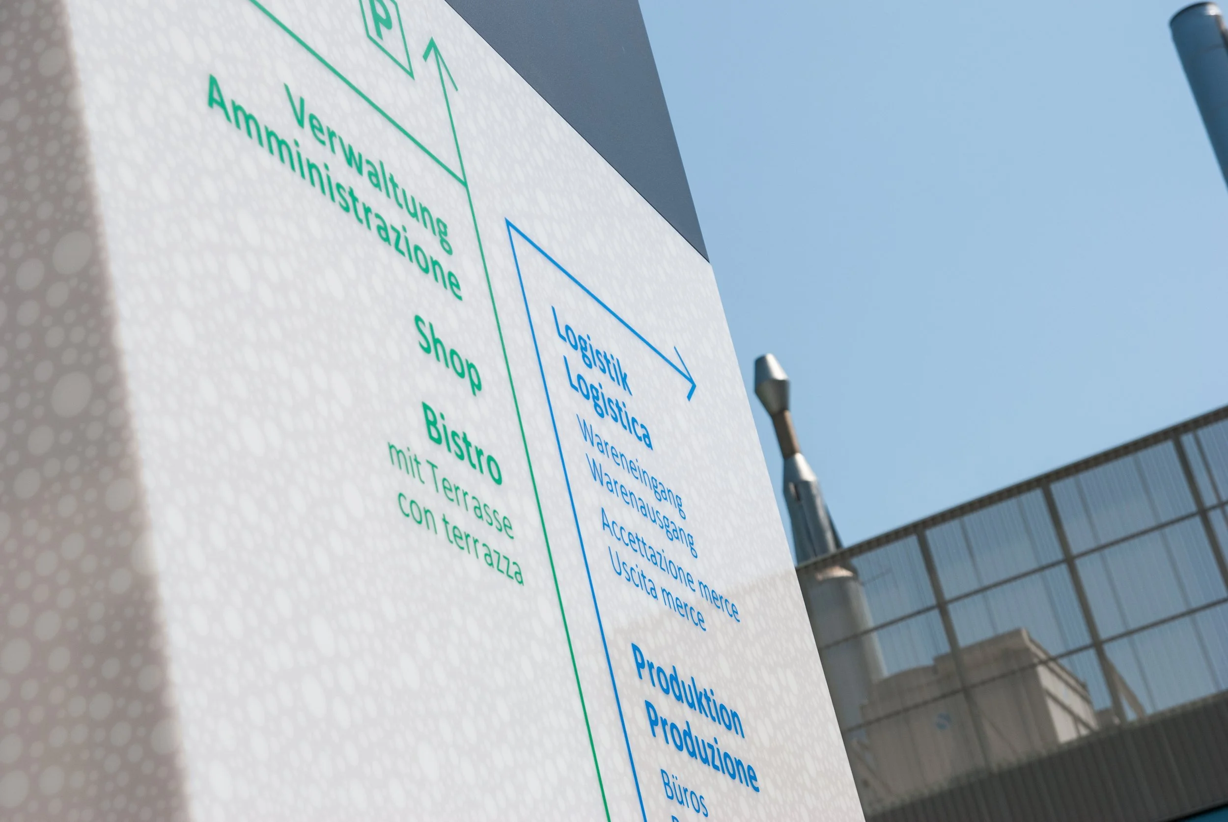 Hinweisschild mit grünen und blauen Texten in deutscher und italienischer Sprache auf dem Gelände einer Molkerei, im Hintergrund ist ein Gebäude und der blauer Himmel sichtbar.