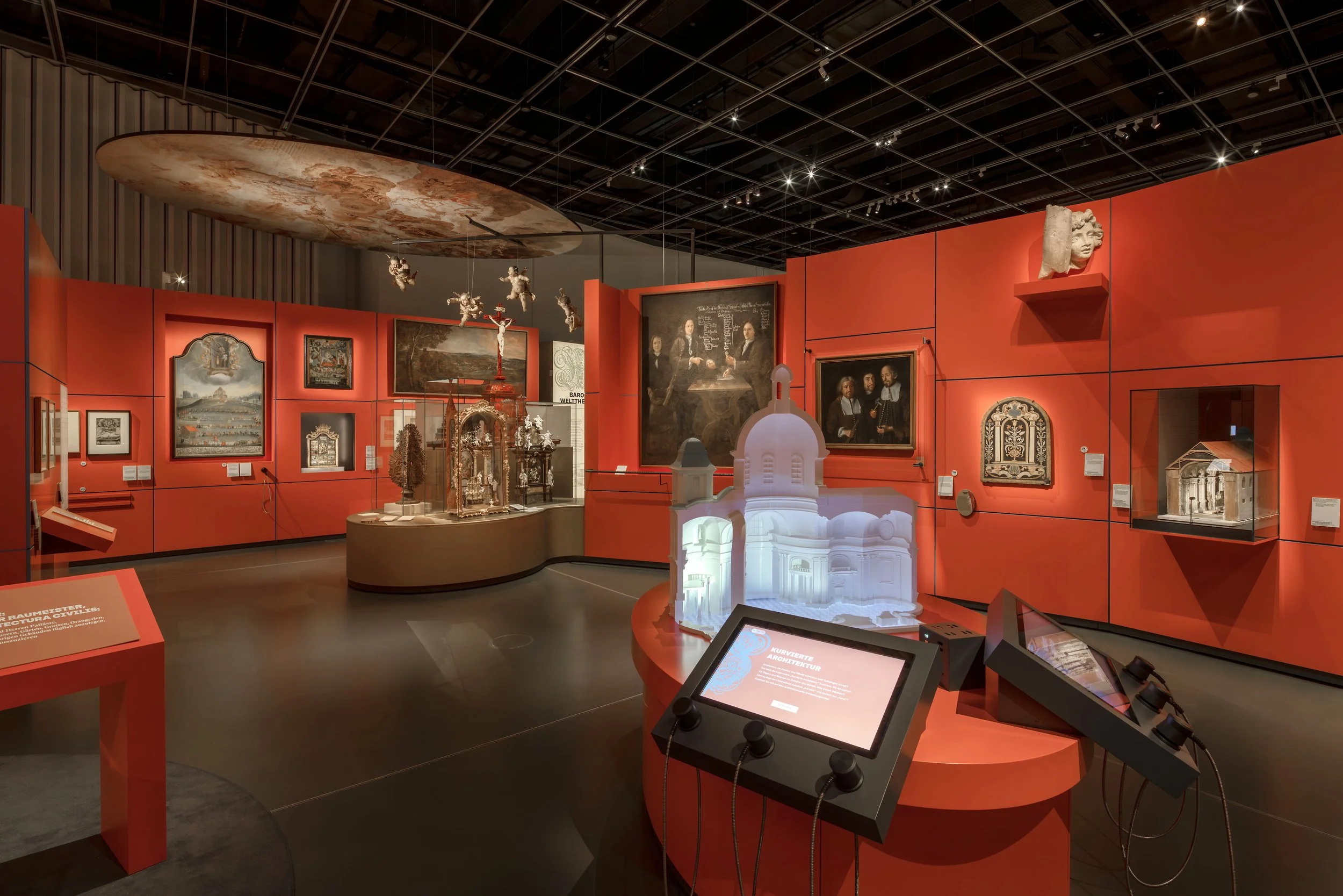 Ausstellung im Museum mit Bildern, Skulpturen und historischen Artefakten an orangefarbenen Wänden, beleuchtete Vitrinen und interaktive Displays.