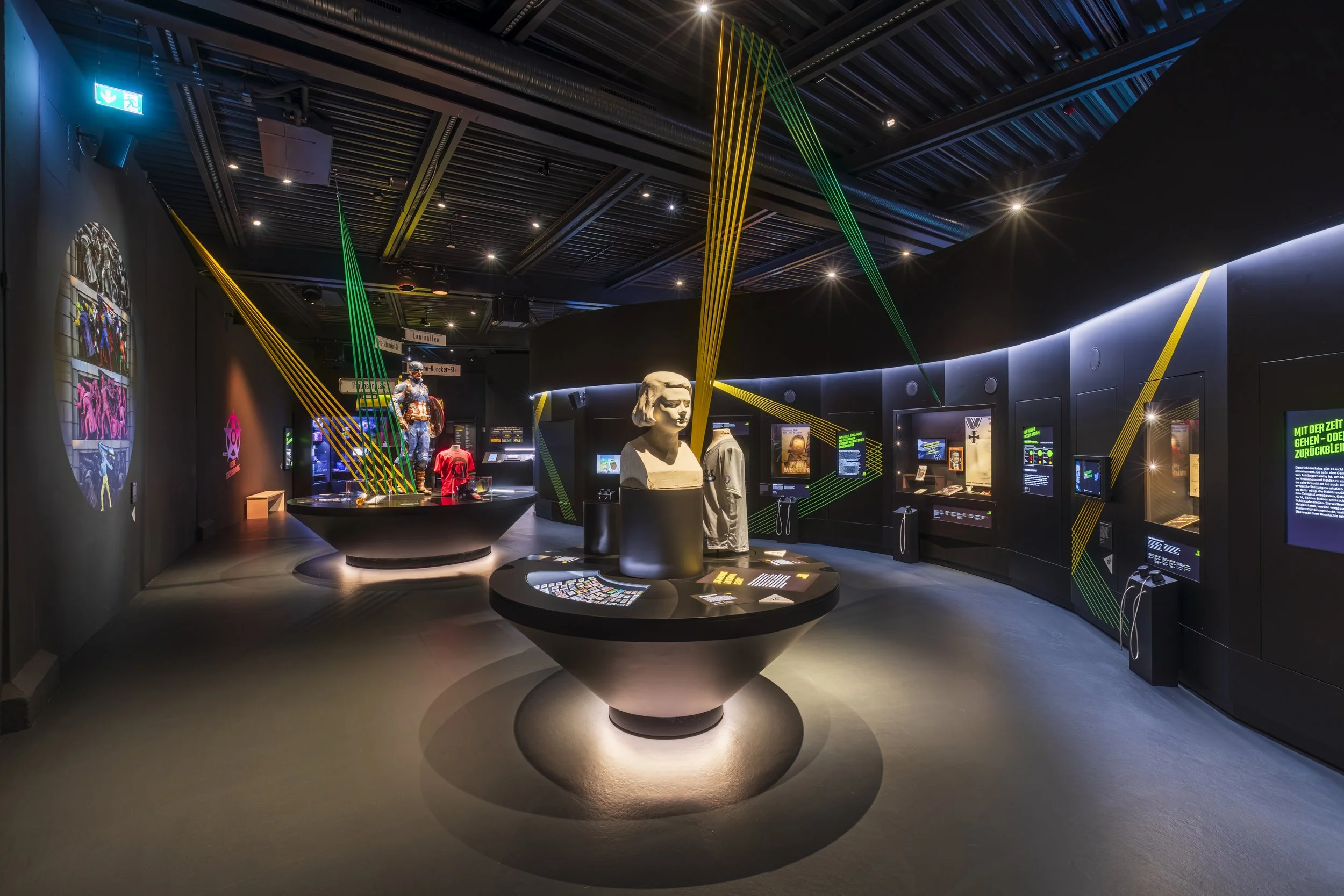 Ausstellungsraum in einem Museum mit dunklen Wänden, erzählerischen Objekten wie Statuen, Kleidung und Digitaldisplays, beleuchtet mit modernem Lichtdesign.