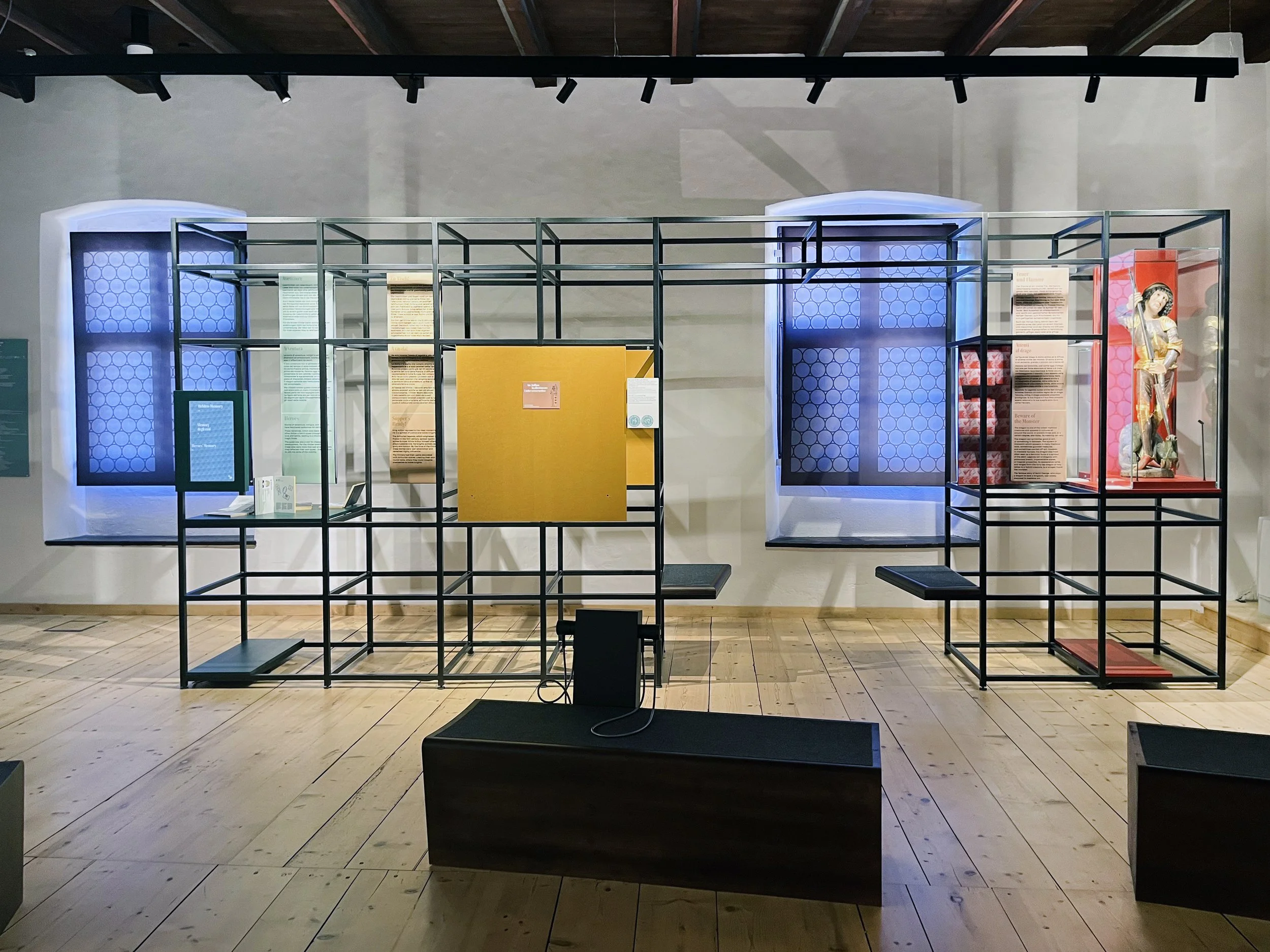 Ausstellung in Schloss Runkelstein mit Sprossenregalen, Texttafeln, einer Statue und blauen Hintergrundleuchten an den Fenstern.