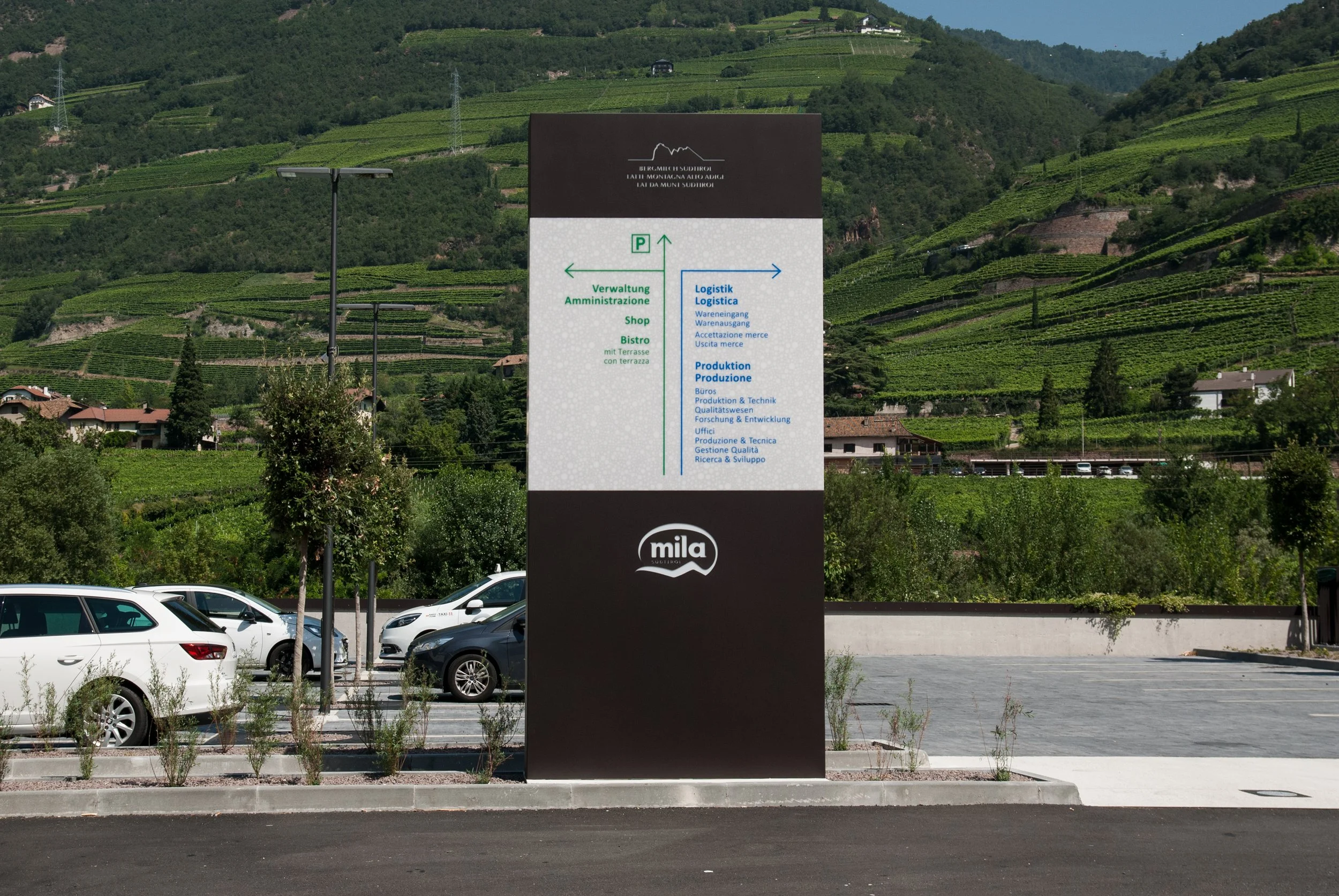 Informationsschild auf einem Parkplatz mit grünen Hügeln im Hintergrund, zeigt Richtungen für Verwaltung, Shop, Bistro, Logistik und Produktion.