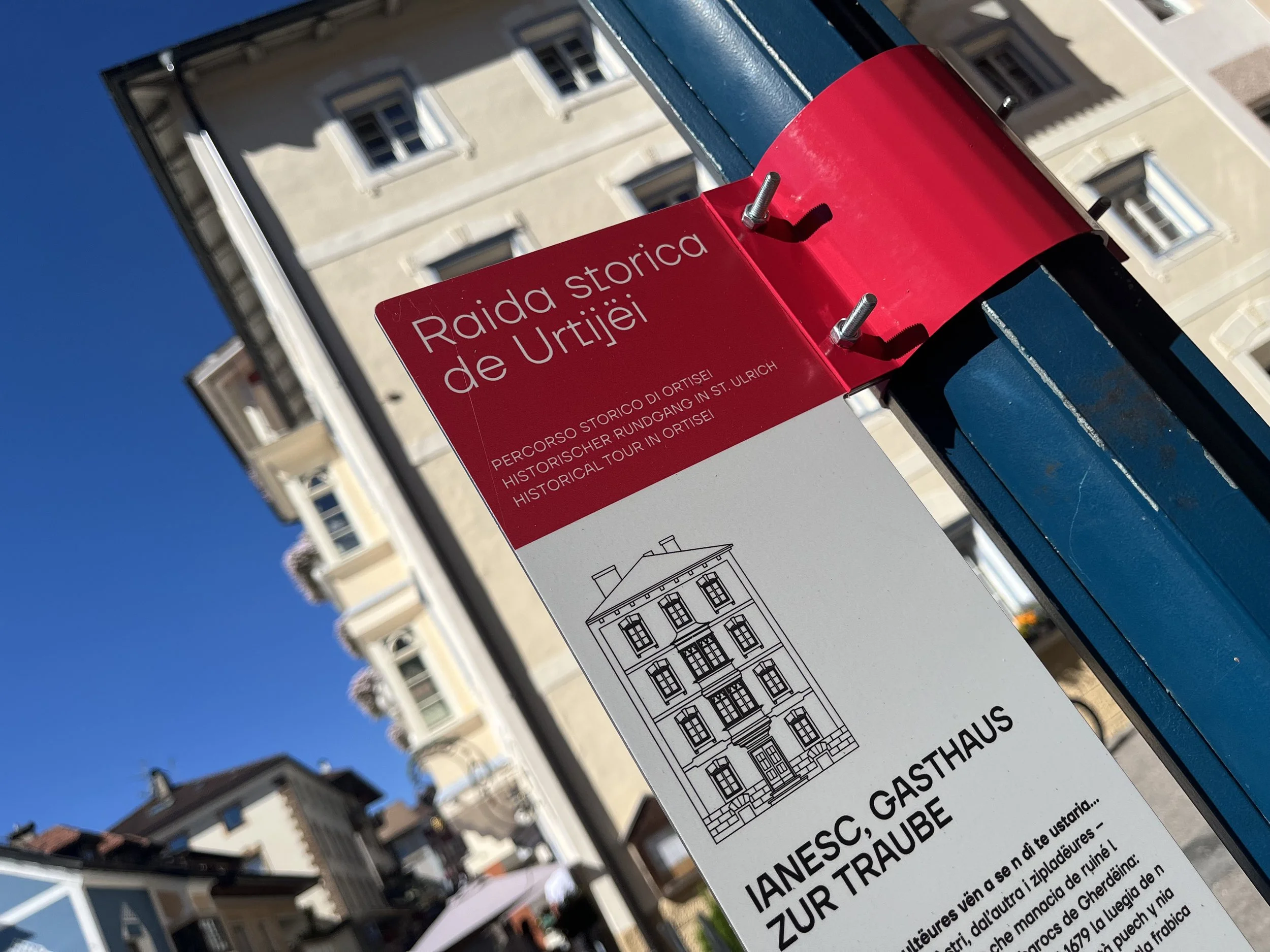 Hinweisschild für eine historische Tour in Ortisei, mit einer Illustration eines Gebäudes und Text in Italienisch, Deutsch und Englisch am Rathaus.