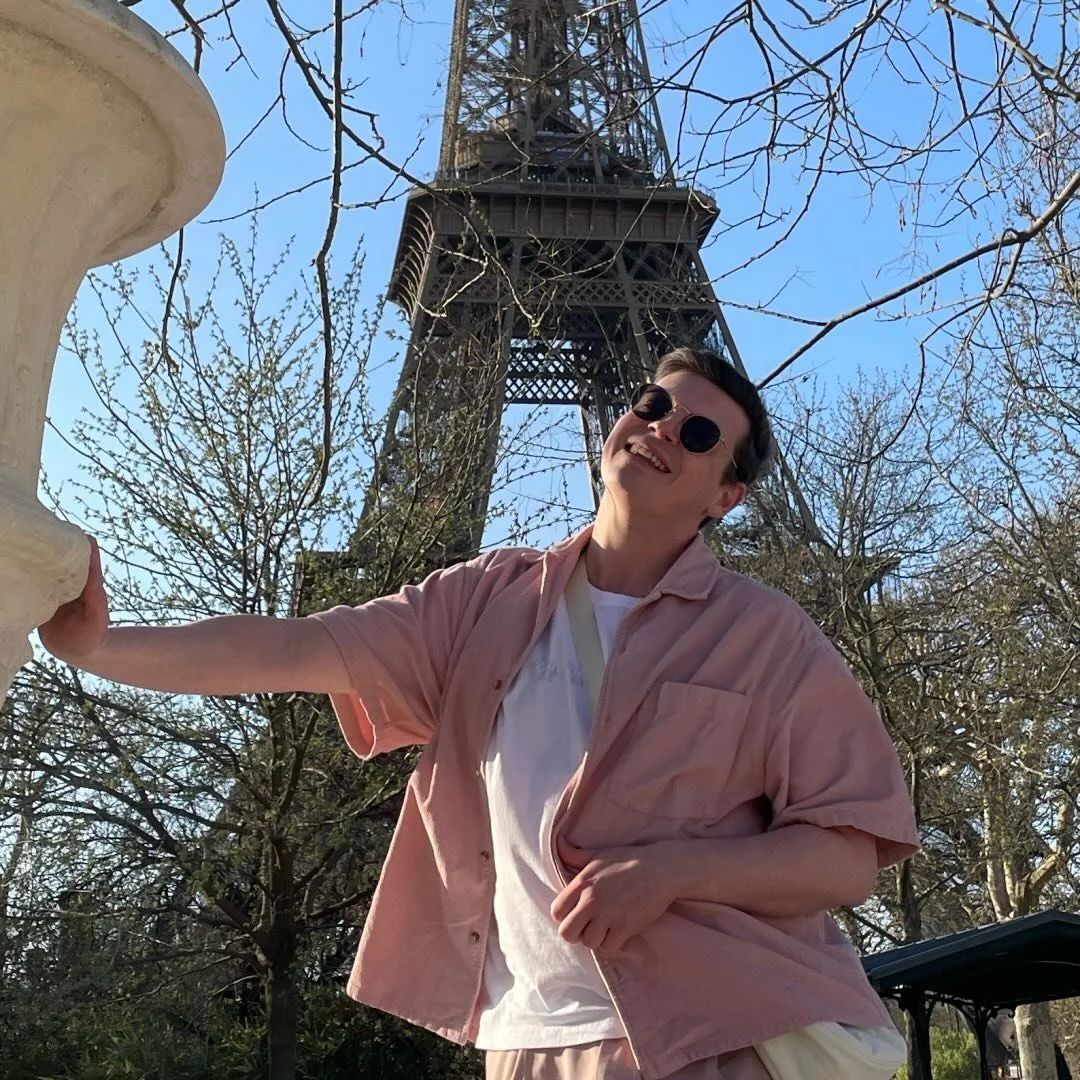 A person smiling and wearing sunglasses, standing outdoors near the Eiffel Tower, with trees and a clear blue sky in the background.