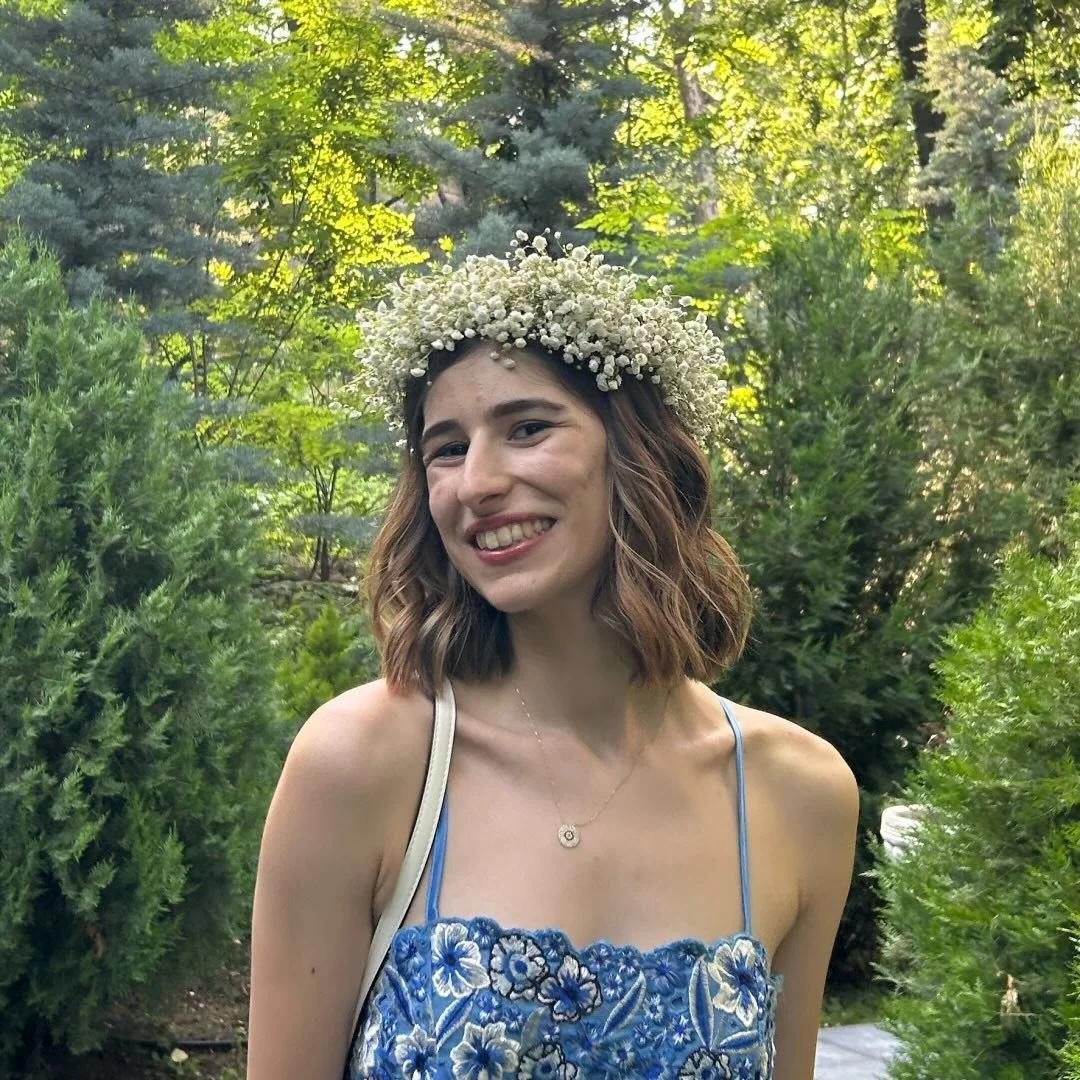 A young woman smiling outdoors in a lush green garden, wearing a blue embroidered dress and a flower crown made of white baby's breath flowers.