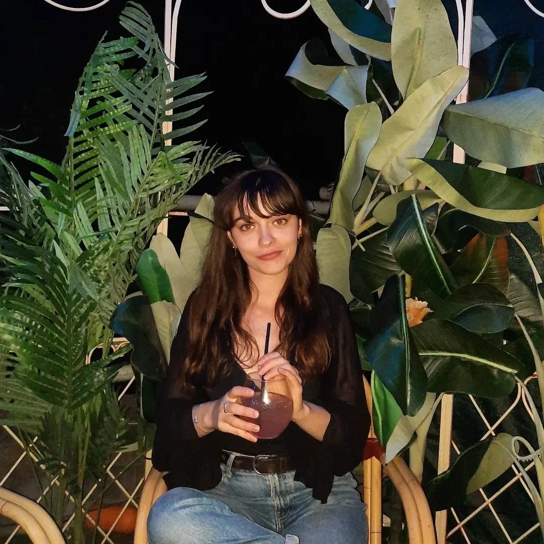 A young woman with long brown hair and bangs, wearing a black top and jeans, sitting outdoors surrounded by lush green tropical plants, holding a dark beverage with a straw.