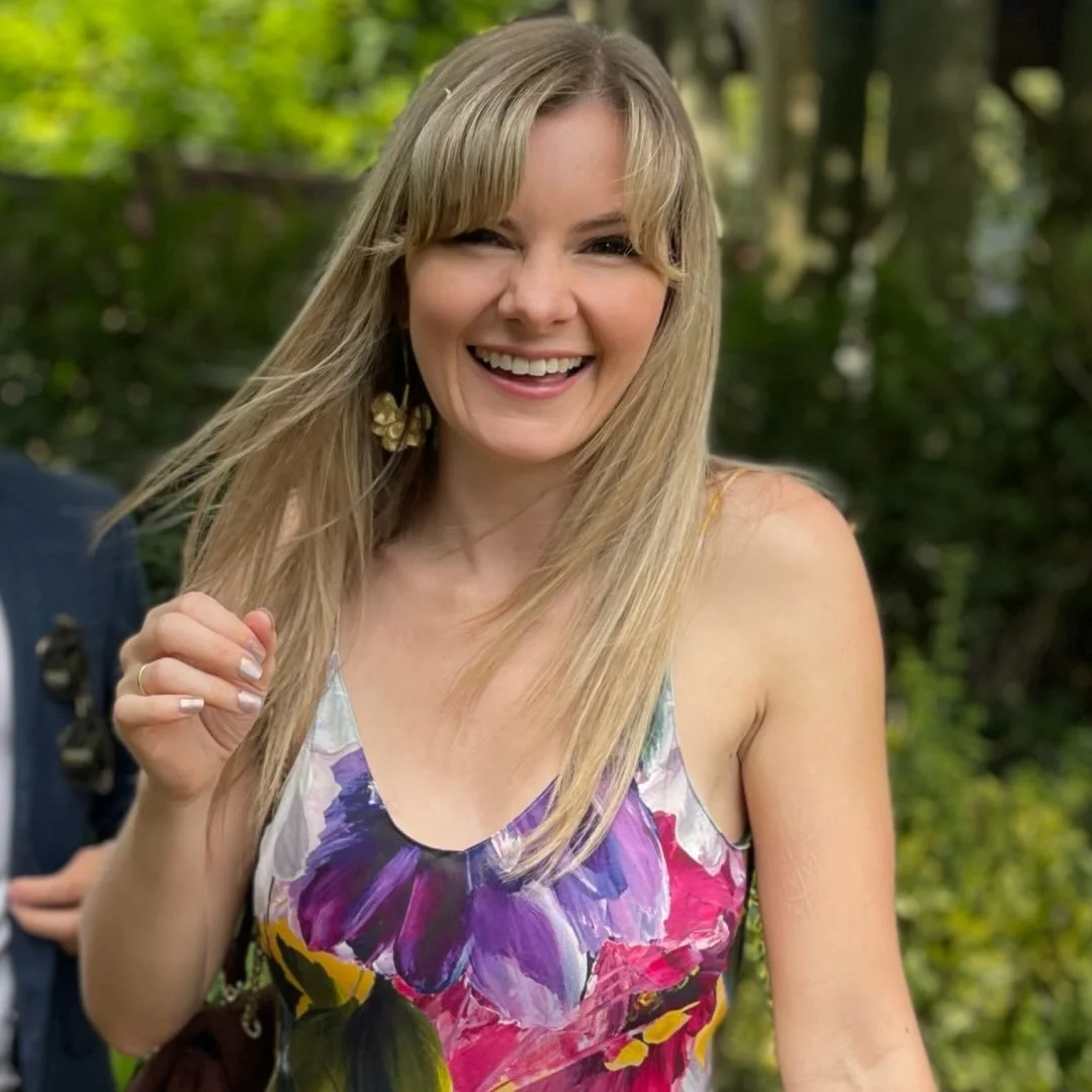 A woman with blonde hair smiling and winking, wearing a floral dress, in an outdoor setting with green trees in the background.