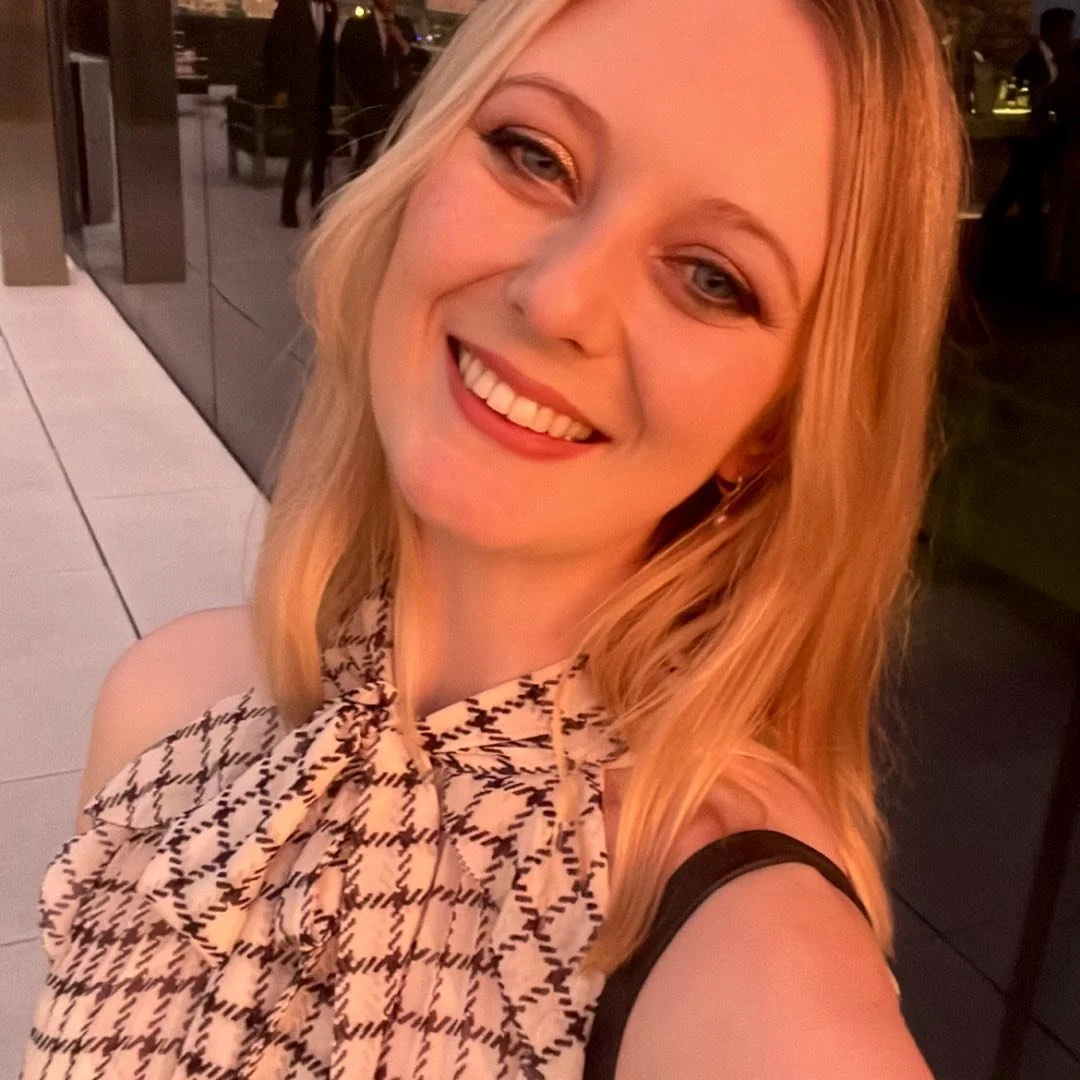 A young woman with blonde hair smiling and taking a selfie at night in an outdoor setting, wearing a patterned top with a bow.