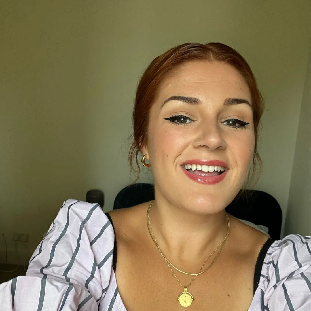 A young woman with red hair taking a selfie, wearing makeup, gold earrings, a nose piercing, layered gold necklaces, and a striped white and black top.