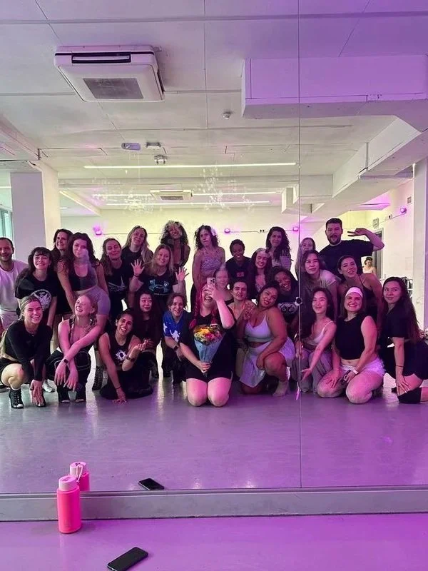 Group of women and one man posing for a photo in a dance studio in Nottingham with purple lighting, mirror, and a concrete ceiling.