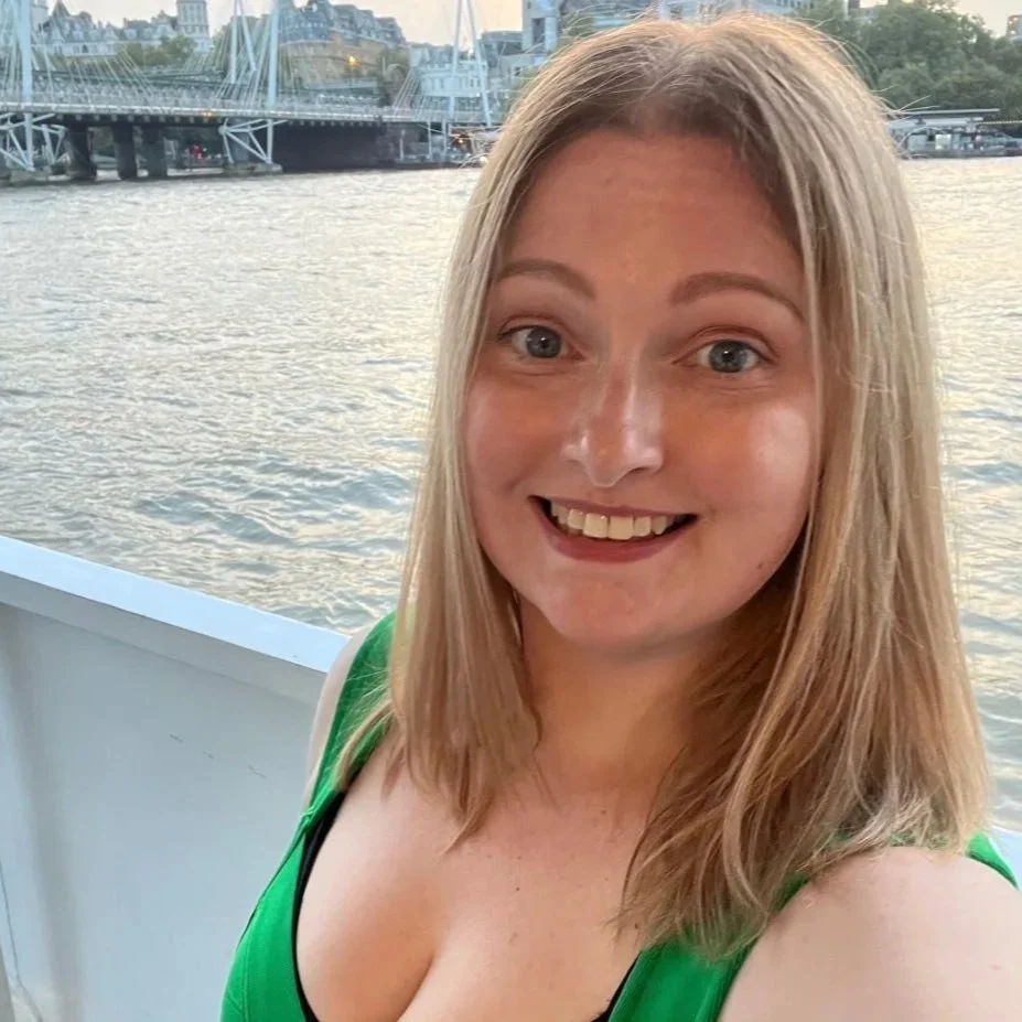 A smiling woman with blonde hair taking a selfie on a boat with a river and a city bridge in the background.