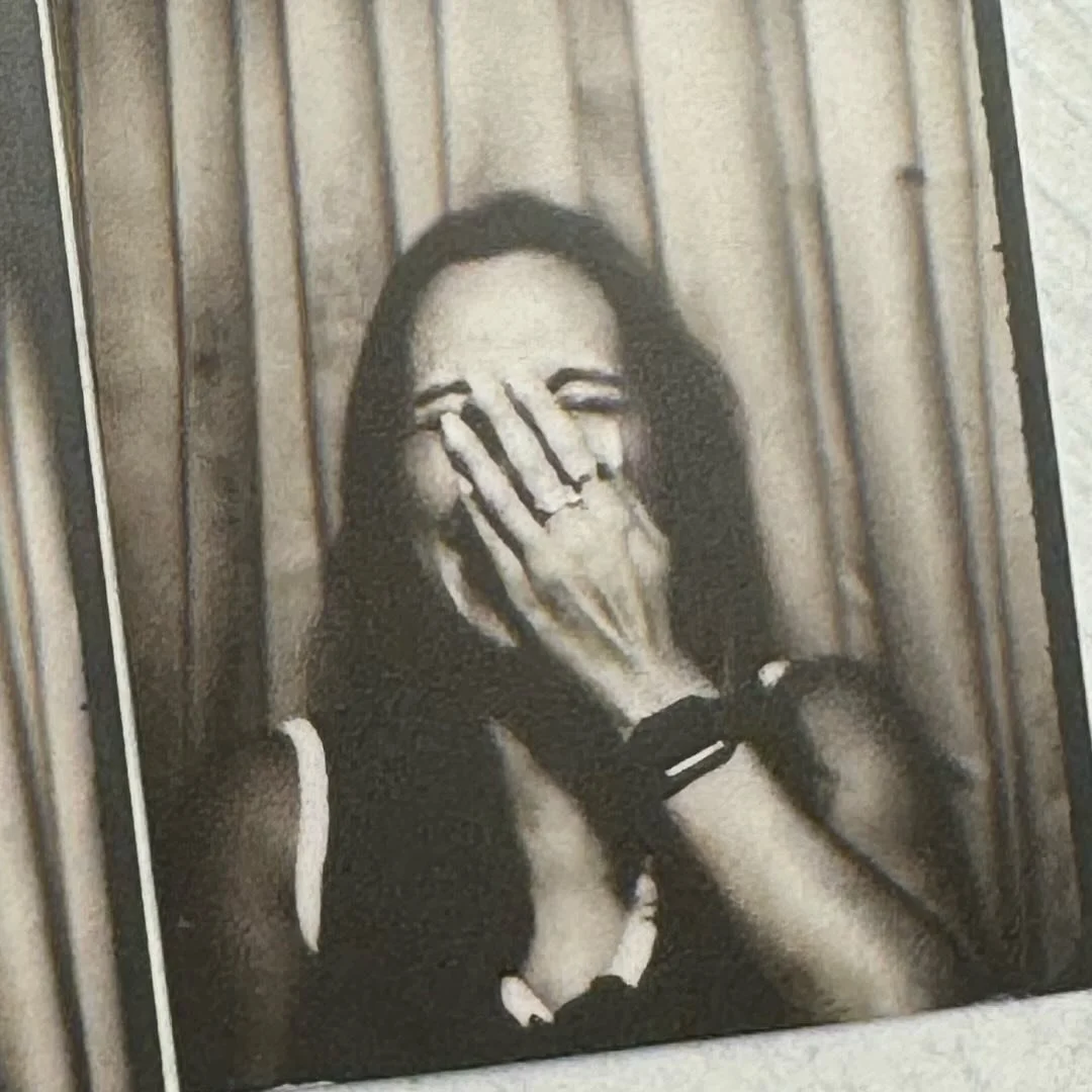 A black and white painting of a young woman covering her face with her hand, standing against a wooden background.