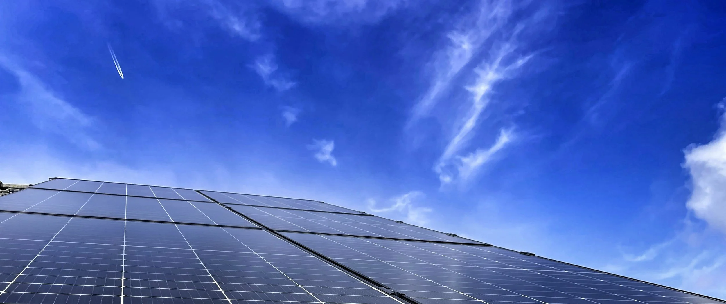 Photovoltaic solar panels installed on a building rooftop against a blue sky with wispy clouds and a jet leaving a contrail.