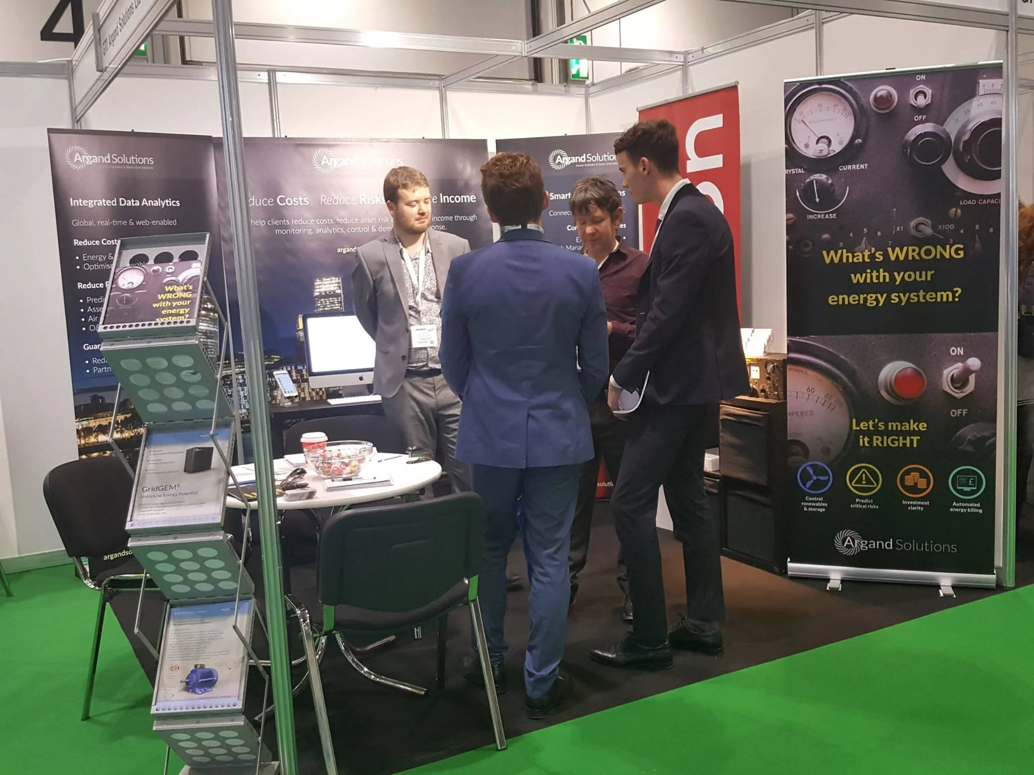 A trade show booth for Argand Solutions with a banner asking 'What's WRONG with your energy system?' and another with technical gauges. Four men are standing and talking inside the booth, with a small table and chairs in front, and brochures displayed on a stand.
