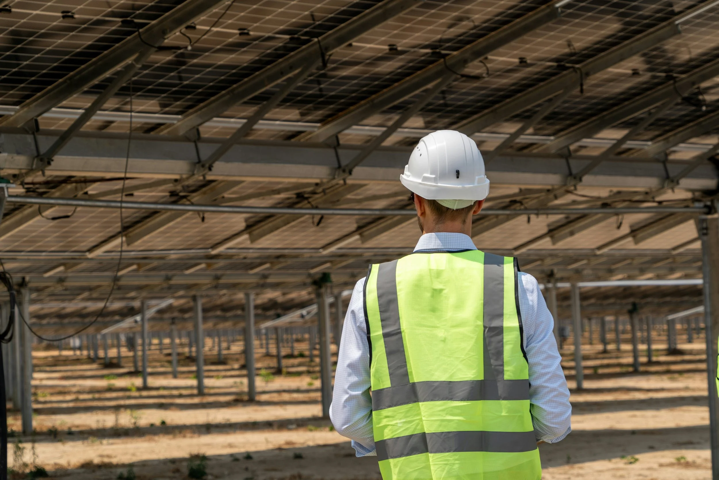 A person in a white hard hat and high-visibility vest inspecting solar panels in a solar farm.