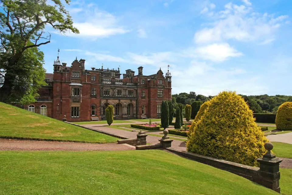 A large, historic mansion with red brick walls, turrets, and decorative architectural details, surrounded by well-maintained gardens with trimmed bushes, pathways, and lawns under a partly cloudy sky.