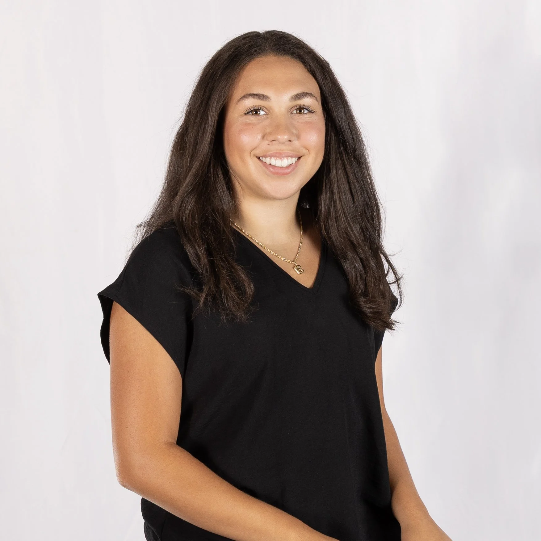 A young woman with long brown hair, wearing a black top and a gold necklace, smiling at the camera against a light background.
