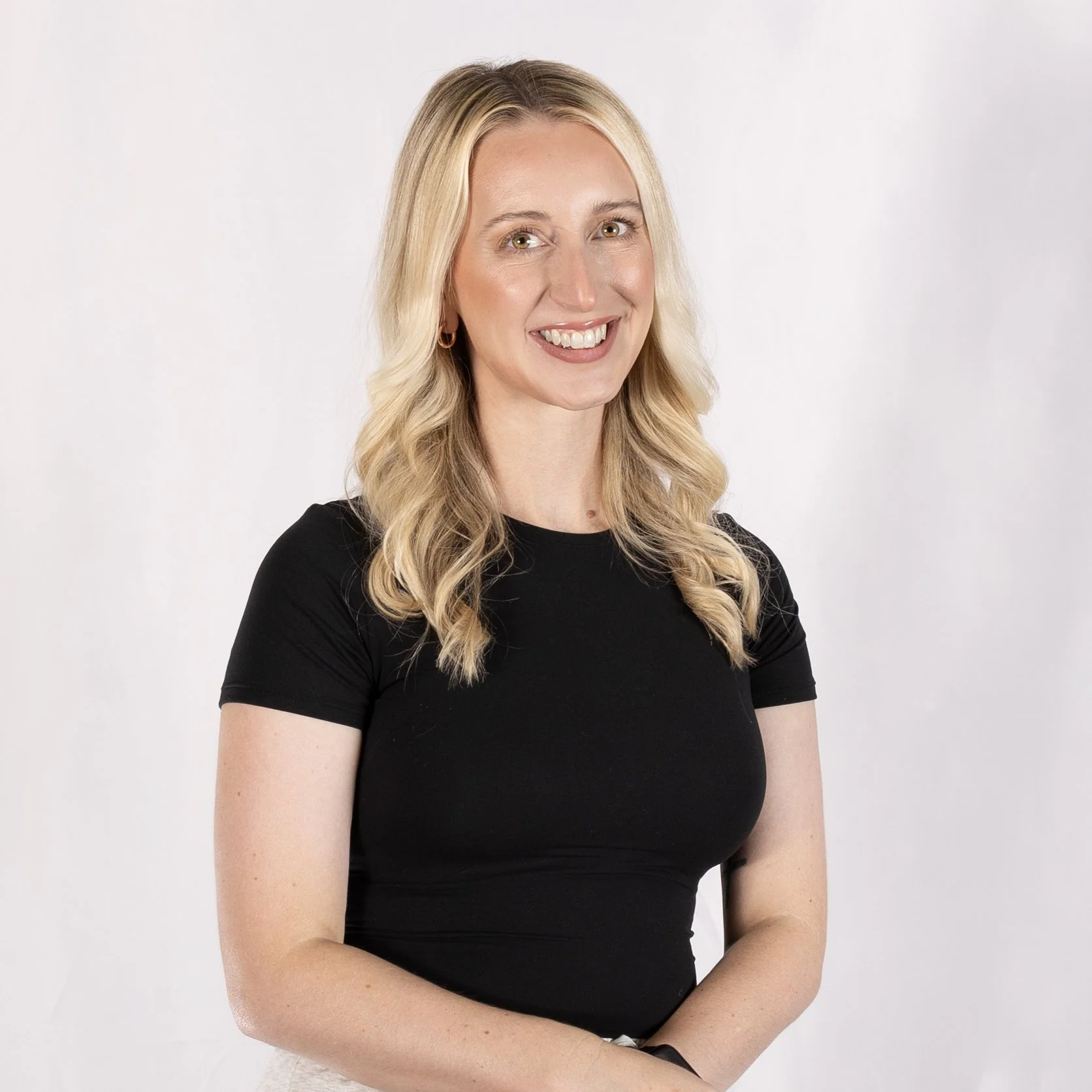 Portrait of a smiling blonde woman with wavy hair, wearing a black t-shirt, against a plain light background.