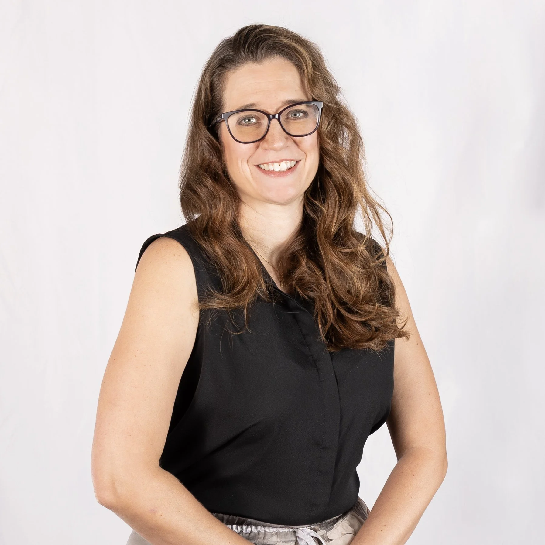A smiling woman with long, wavy brown hair, wearing glasses, a sleeveless black top, and patterned pants against a plain white background.