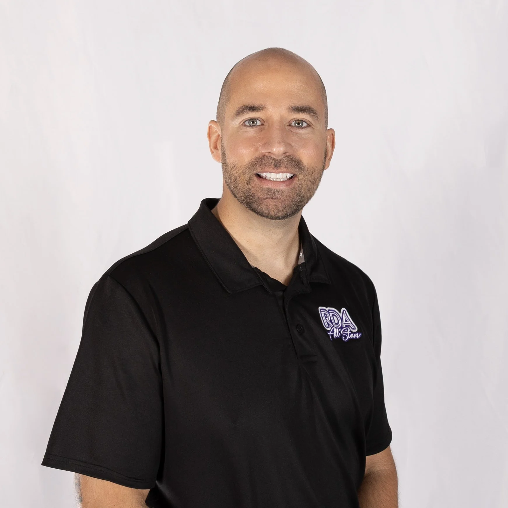 A man with a shaved head and beard, smiling, wearing a black polo shirt with a logo on the chest that reads "RDA All Star," against a plain white background.