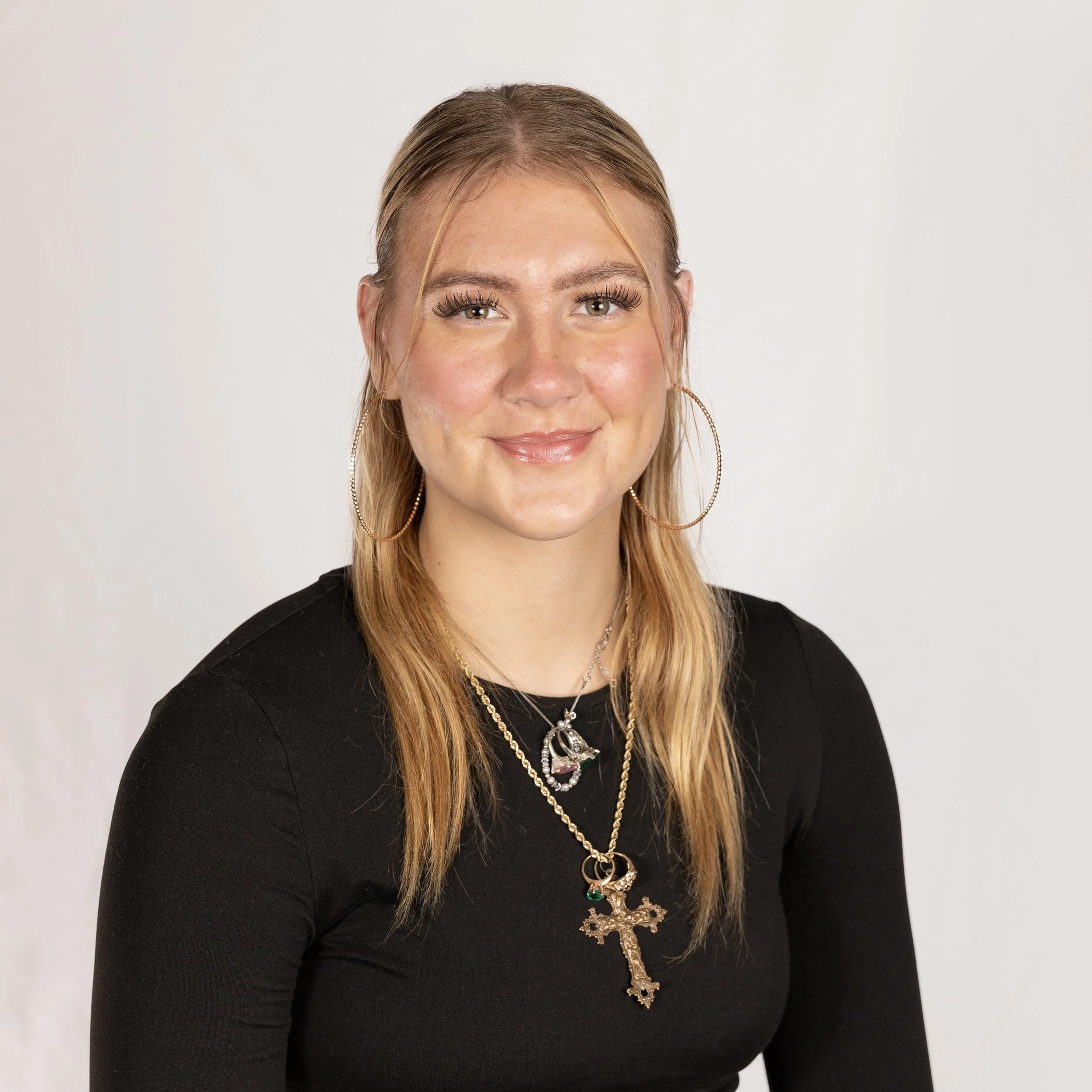A young woman with long blonde hair, wearing a black top, gold hoop earrings, layered necklaces with pendant charms, and smiling at the camera with a neutral background.