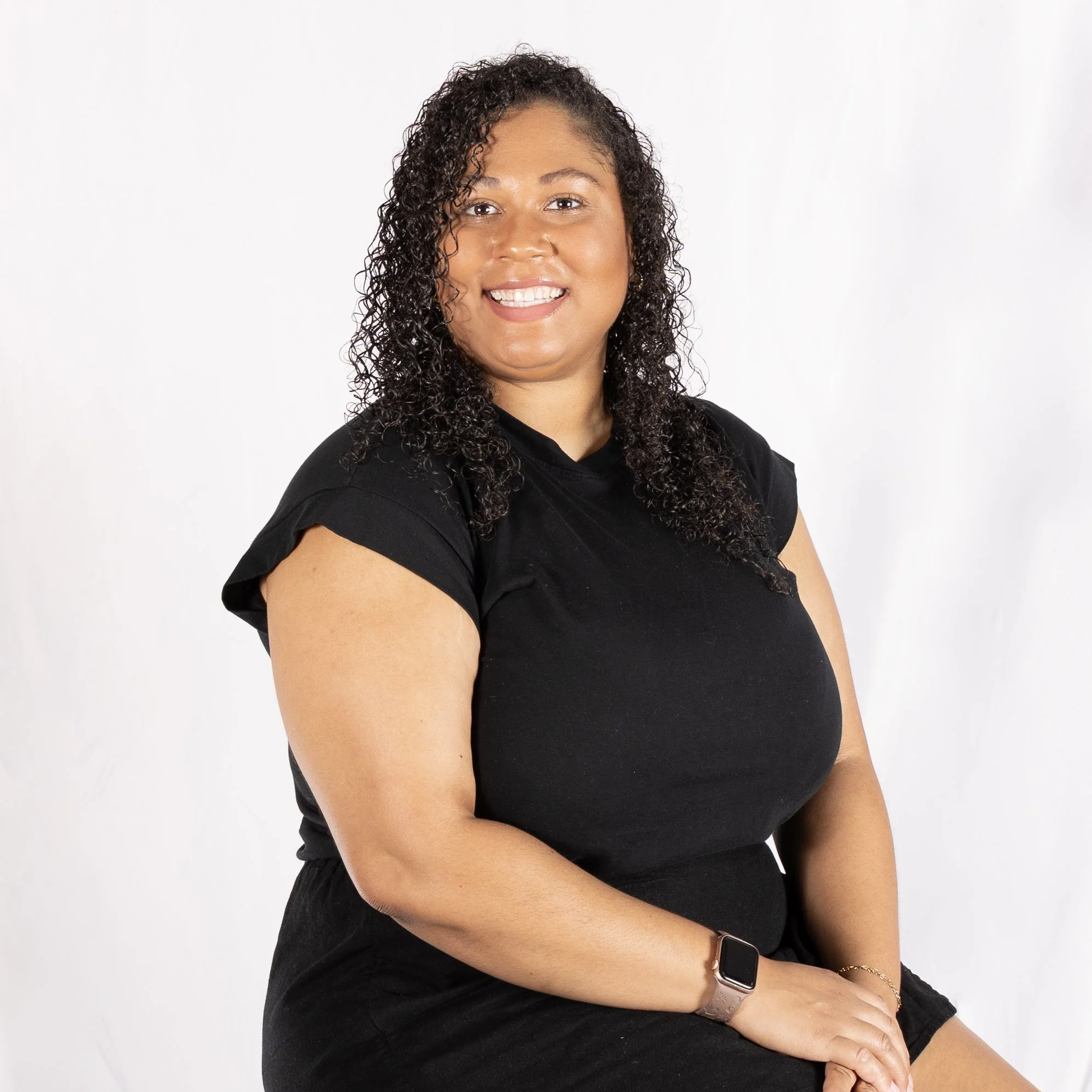 A woman with curly dark hair wearing a black t-shirt and shorts, smiling against a plain white background.