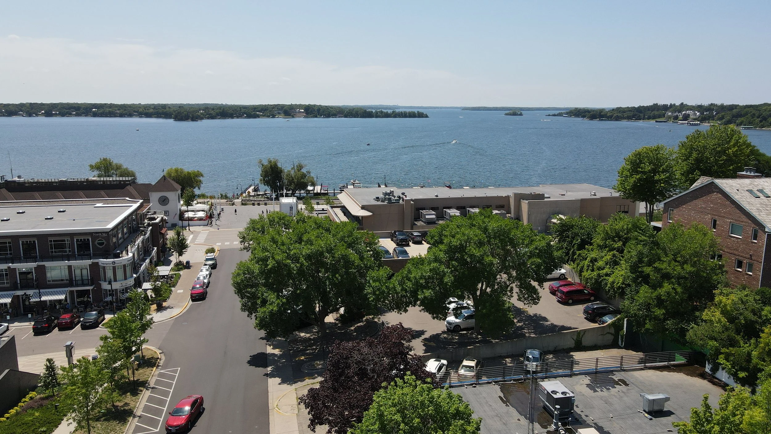 Aerial view of downtown Wayzata, Minnesota, near Lake Minnetonka & Wayzata Bay with boats, buildings, trees, and a parking lot.