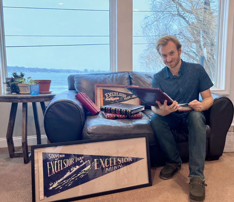 A man sitting on a gray couch in a bright room with large windows, holding a book and smiling at the camera. On the floor in front of him is a framed souvenir sign that reads 'Souvenir Excelsior Minn.' To his left, there are stacked books and a framed picture with 'Excelsior Park' on it. A small table to the side holds potted plants and a blue cup.