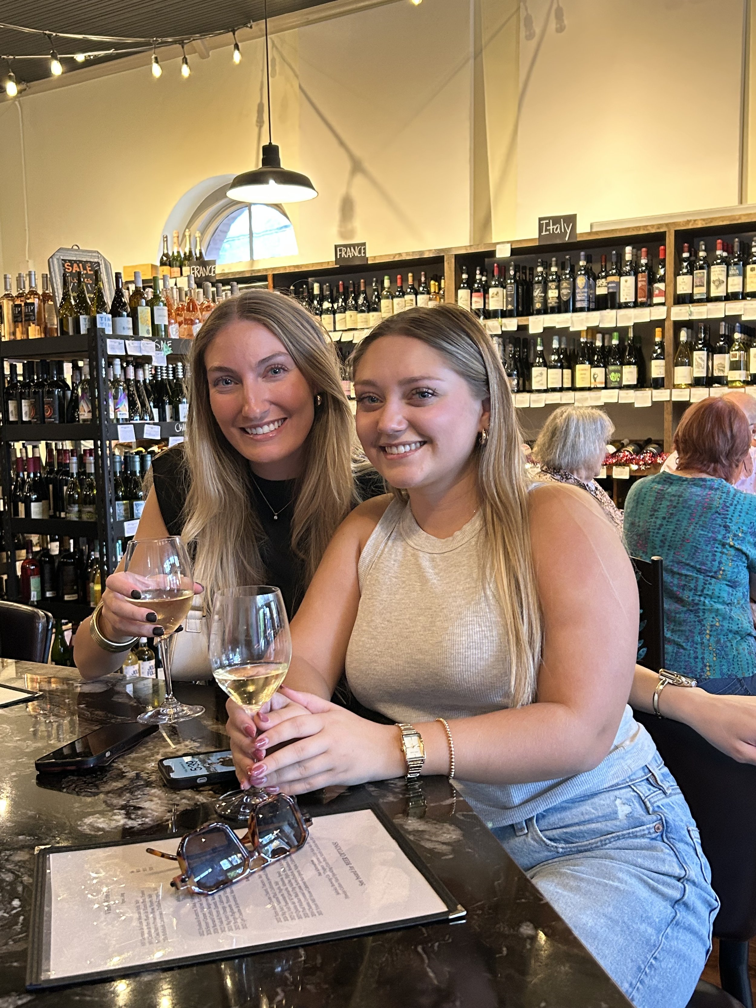 Two women sitting at a table inside a wine bar, smiling and holding glasses of white wine. Behind them are shelves filled with bottles of wine and a sign indicating different countries, such as France and Italy.