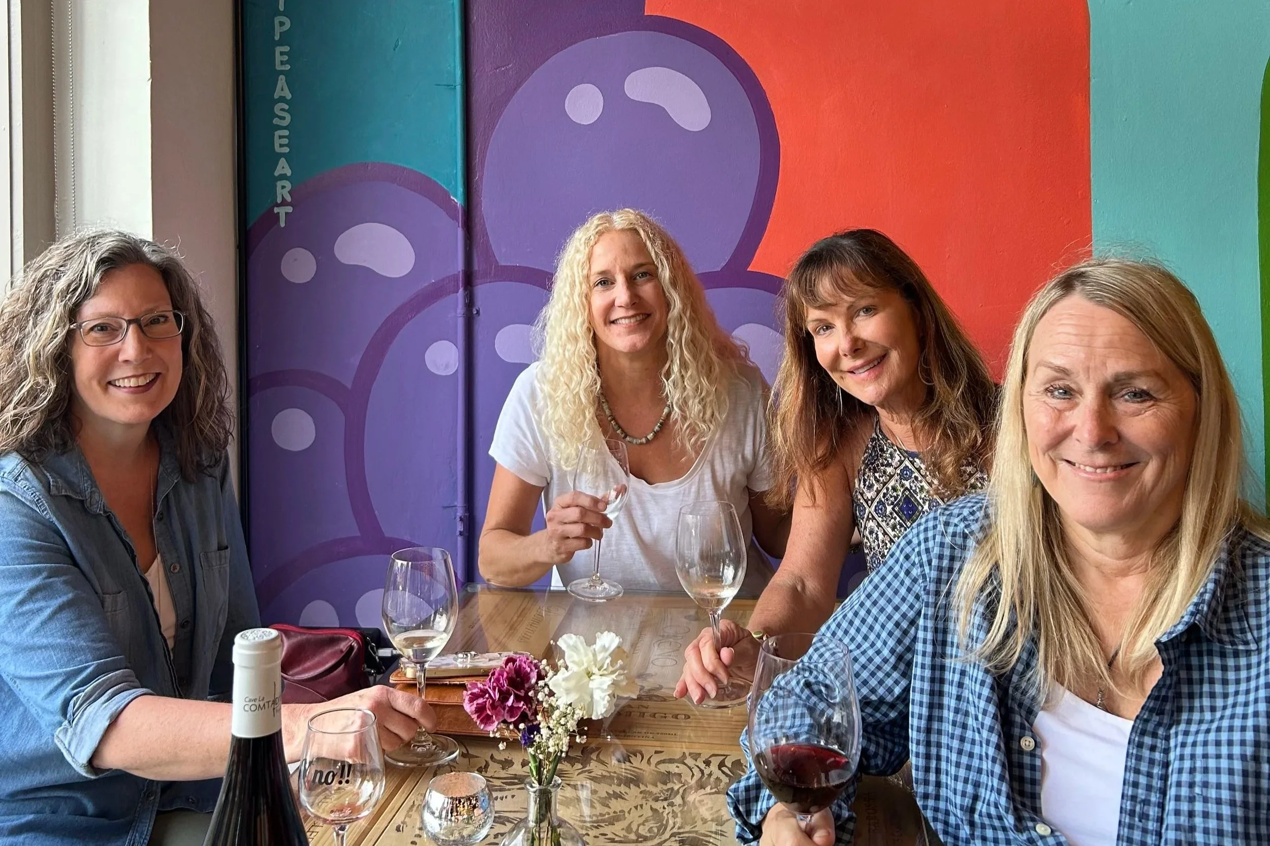 Four women sitting at a table enjoying wine and smiling at a restaurant with colorful purple and orange mural art in the background.